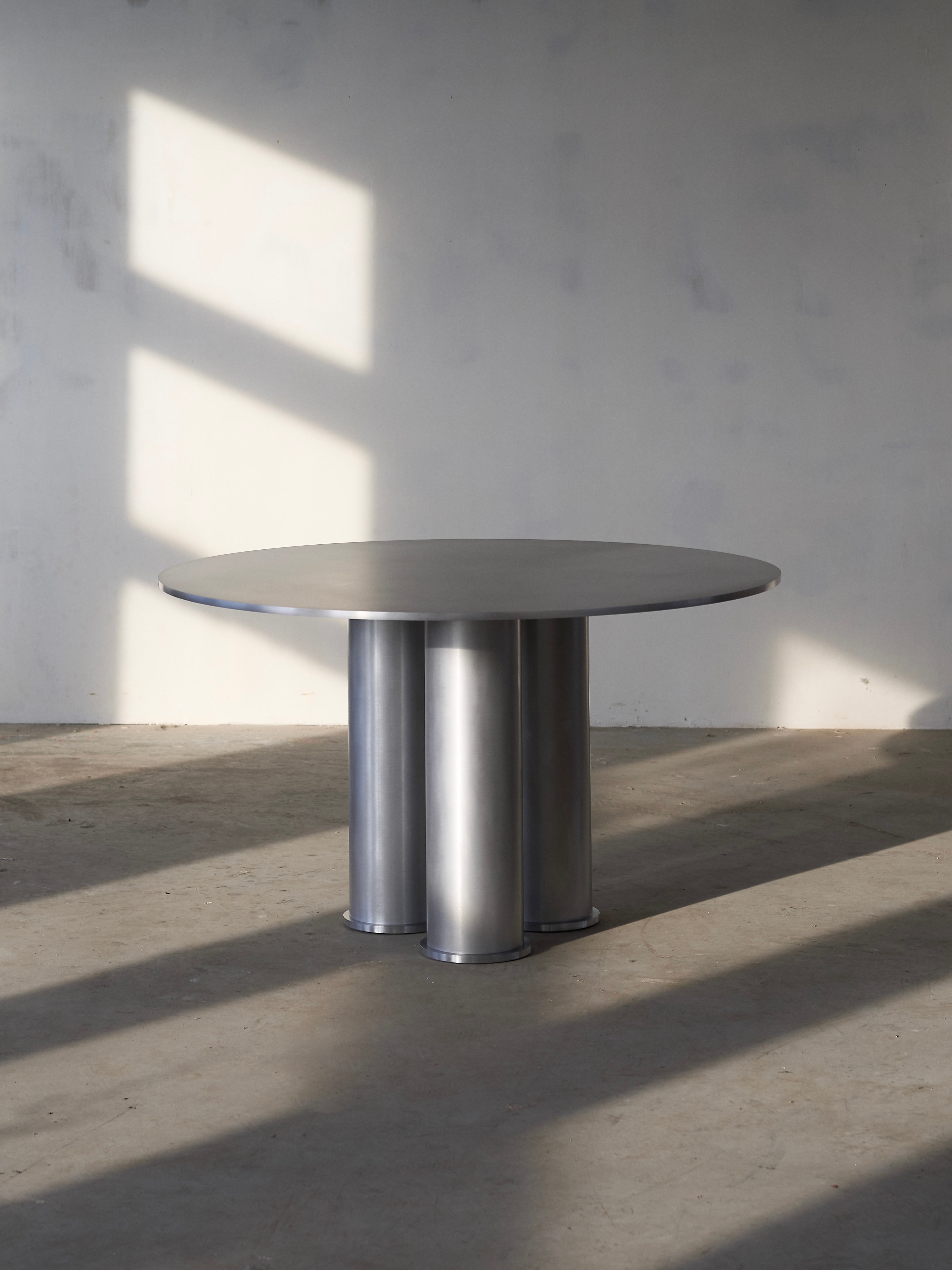 A round, modern Elephant Tripod Table by Frederik Fialin with a metallic finish sits on a bare concrete floor in a minimalist room. Sunlight casts geometric shadows on the wall, emphasizing the table's sleek design and cylindrical base.