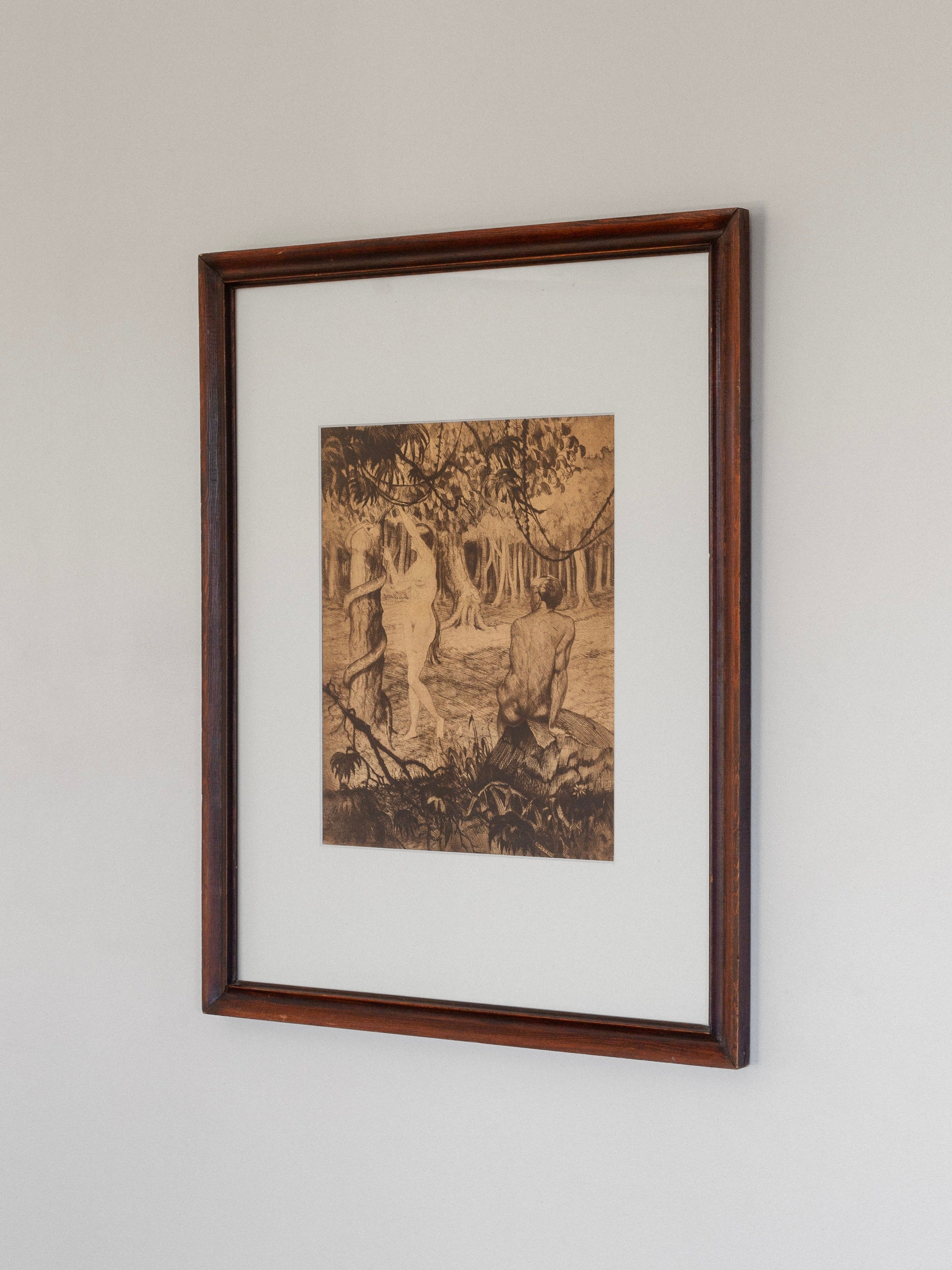 A framed etching titled "Adam & Eve" by Gaston Smidt, created in the 1900s using dry-needle technique, showcases nature with trees and shoreline. Part of Spigel's collection, it features a sepia tone in a dark rustic wooden frame against a light backdrop.