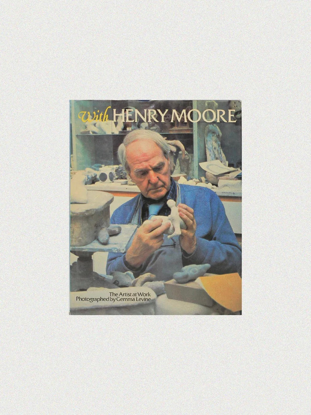 The cover of the Art Book "With Henry Moore" by Boga Avante Shop shows an elderly man in a blue shirt immersed in modern sculpture amidst tools and clay in a cluttered studio. Photo by Gemma Levine.