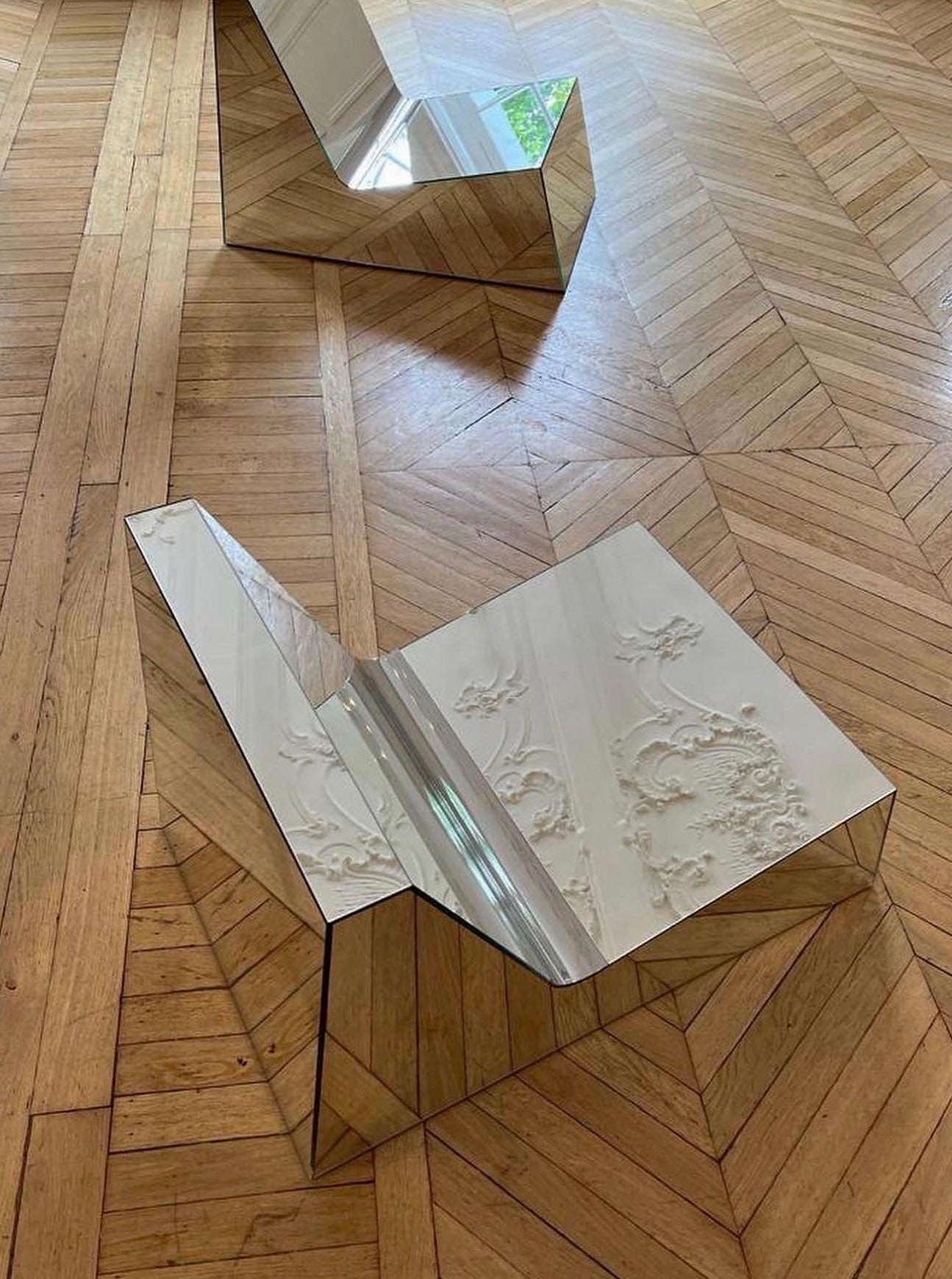 A **Mirror Lounge Chair** by **Project 213A** made of reflective material with intricate patterns on the seat's surface rests on a polished wooden floor with a geometric design. The chair's angular form contrasts sharply with the vintage look of the floor. Another similar chair with a mirror surface is in the background.