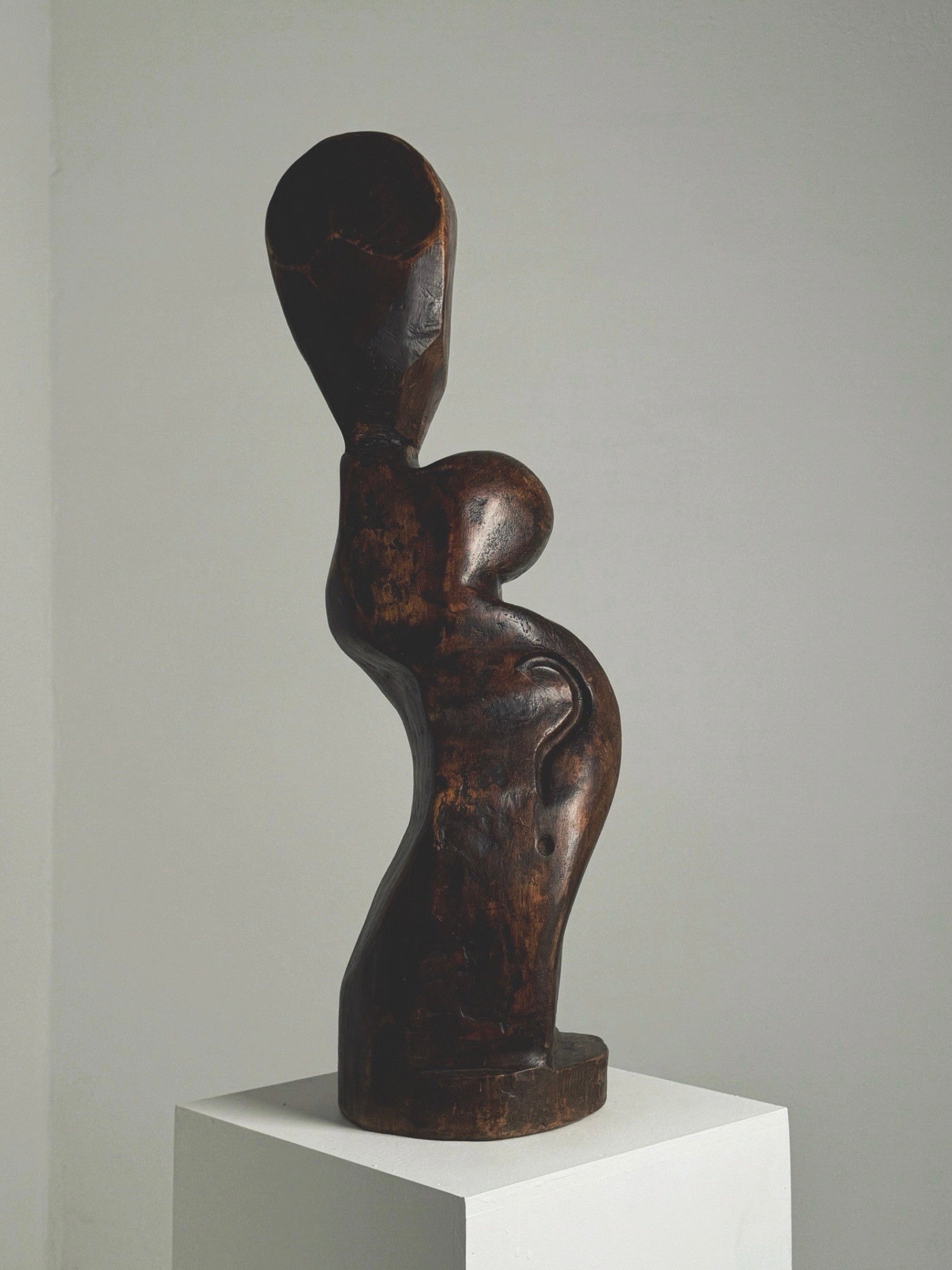 The Large Anthropomorphic Wood Sculpture by Figuresdesign, a dark wooden piece featuring an abstract human shape with a rounded form and elongated head, stands gracefully on a white pedestal against a plain gray background.