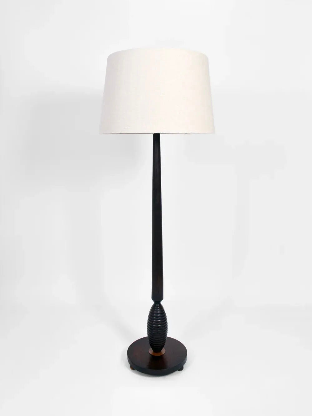 Introducing the Dark Wooden Floor Lamp from Barracuda Interiors, a design that captures the sophistication of 1930s France. This contemporary lamp features a sleek black base complemented by an exquisite textured oval accent. Topped with a large, white fabric drum shade, it stands proudly on a round base, creating an elegant centerpiece against a plain white backdrop.