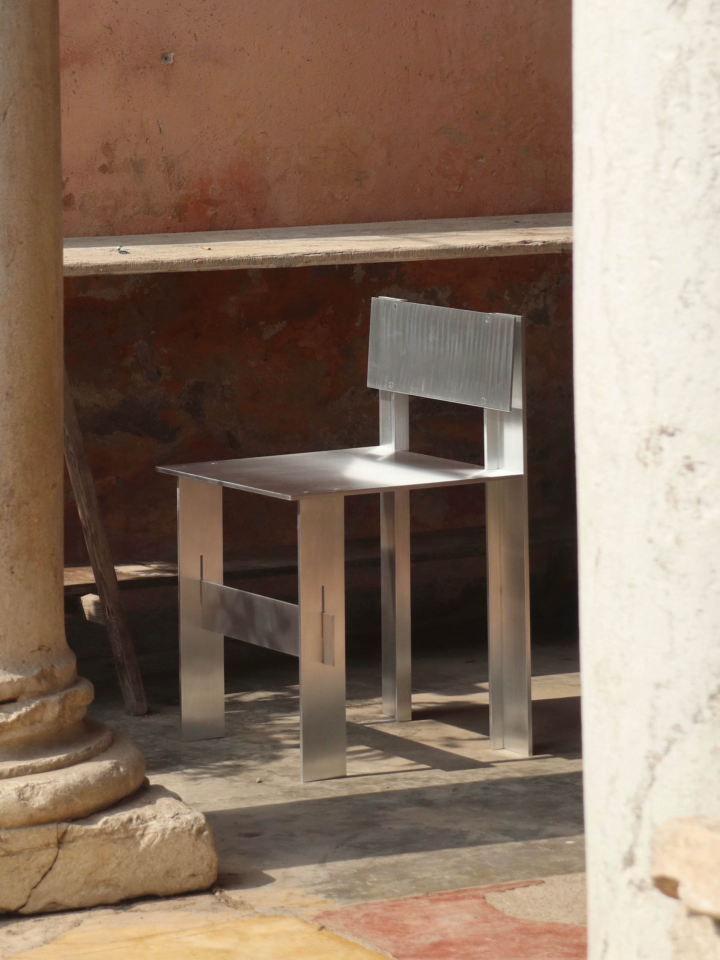 The AL Chair by Filippo Andrighetto sits outdoors on a rustic stone floor amidst ancient columns and a faded pink wall. Soft light highlights its sleek, modern lines, offering a striking contrast to the historical setting.