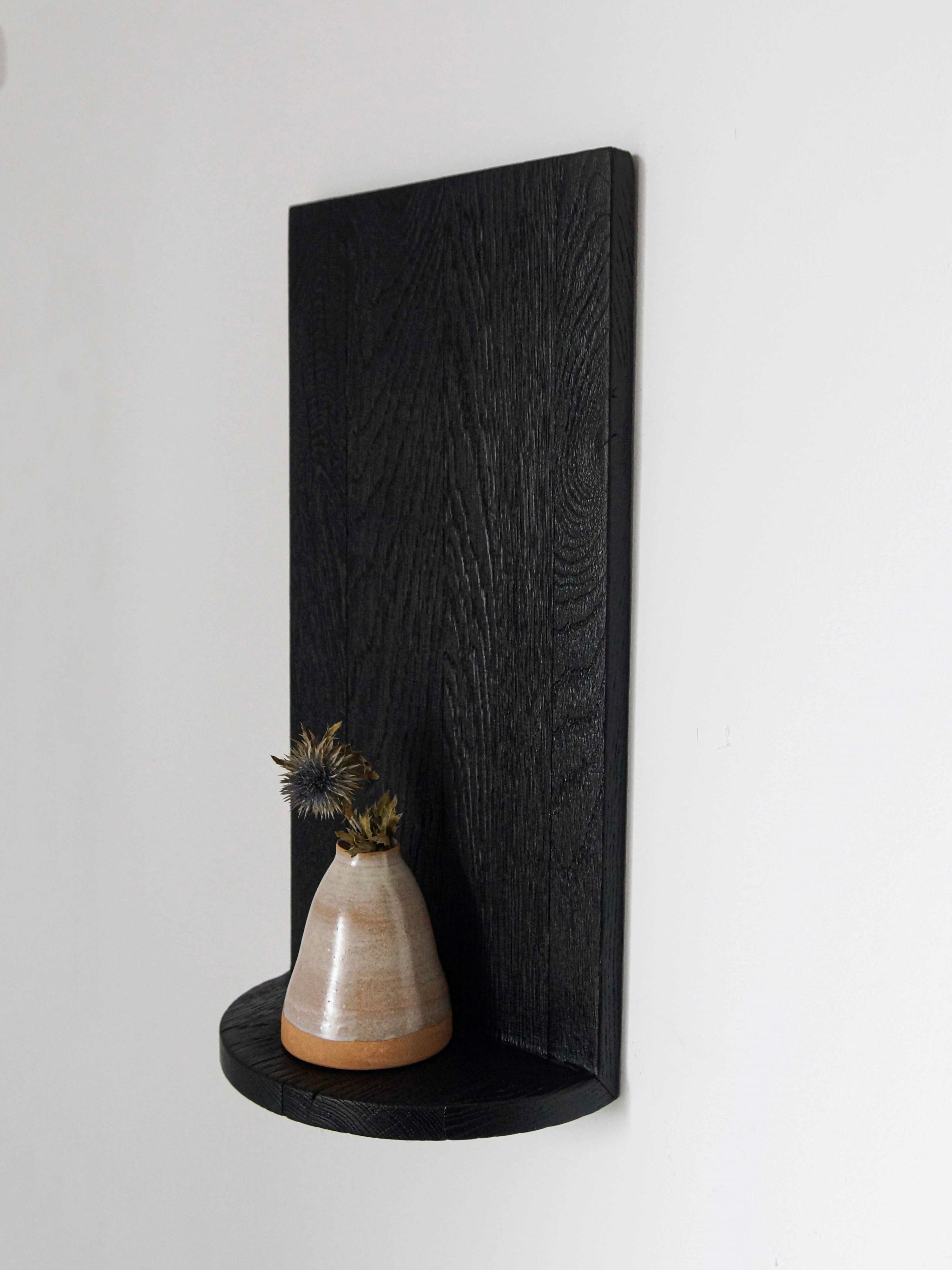 A small ceramic vase with dried thistles sits on the "Megaron" Shelf Black by COSEINCORSO. The minimalist sculptural shelf features burnt natural oak, a vertical back panel, and a small rounded base attached to a white wall, offering a sleek and modern look.
