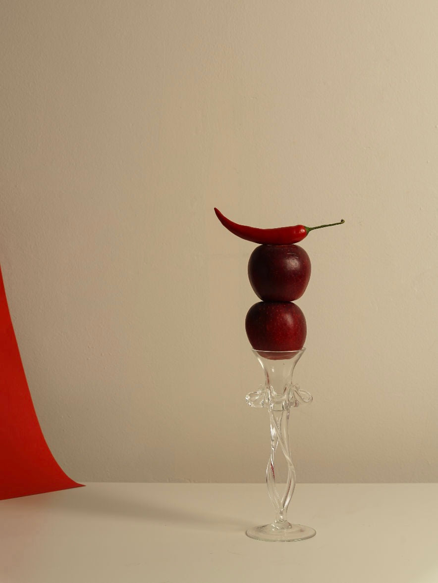 A vertical composition features two red apples stacked on a delicate Mouth Blown Candle Holder by Les Objets Amsterdam. Atop the apples rests a red chili pepper. The minimalistic and artistic still life is enhanced by a neutral background that contrasts with a vibrant red swath of cloth on the left, highlighting the Provençal Prestige collection from Les Objets Amsterdam.
