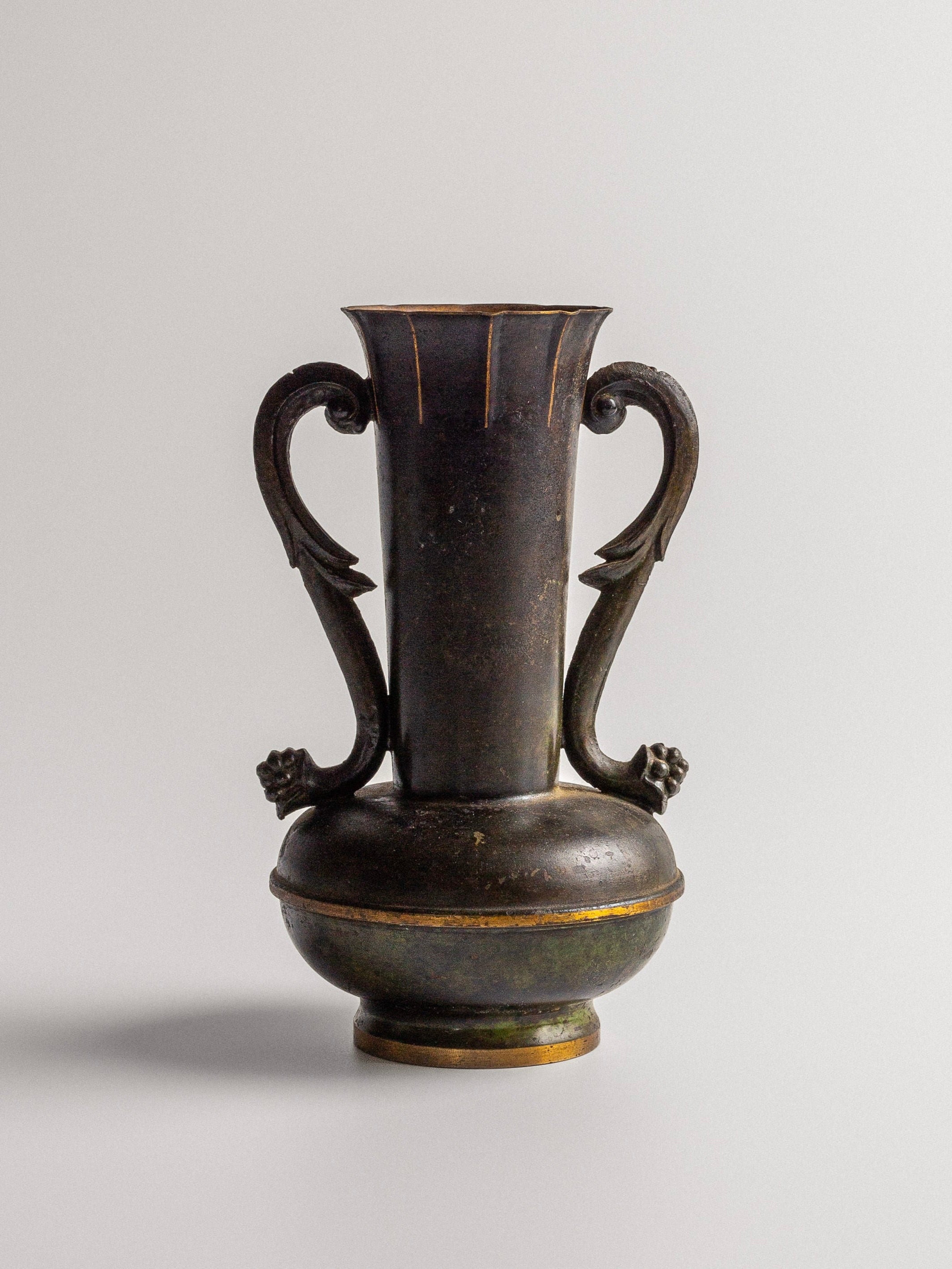 Introducing the Spigel Art Deco Bronze Vase: a dark antique neoclassical masterpiece featuring an elegant design. This metal vase boasts a tall, narrow body with two ornate handles on either side. The patinated bronze finish, round base, and flared rim give it an aged appearance. Displayed against a plain light gray background, this vase exudes timeless sophistication.