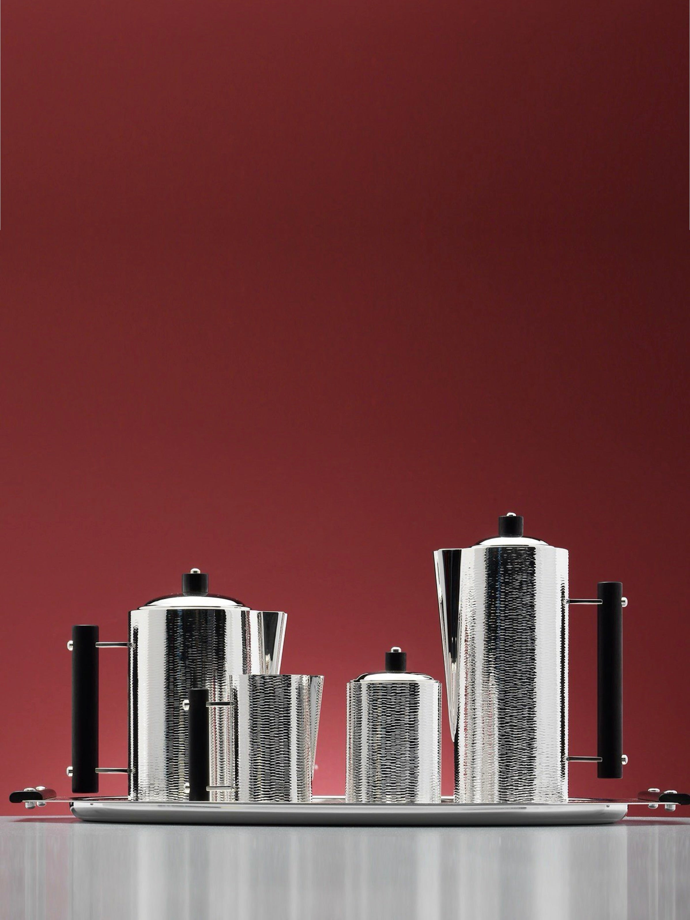 The Ebony Tea Set by zanetto features textured, hand-hammered silver alloy with black handles and cylindrical forms, elegantly displayed on a polished tray against a solid maroon background.