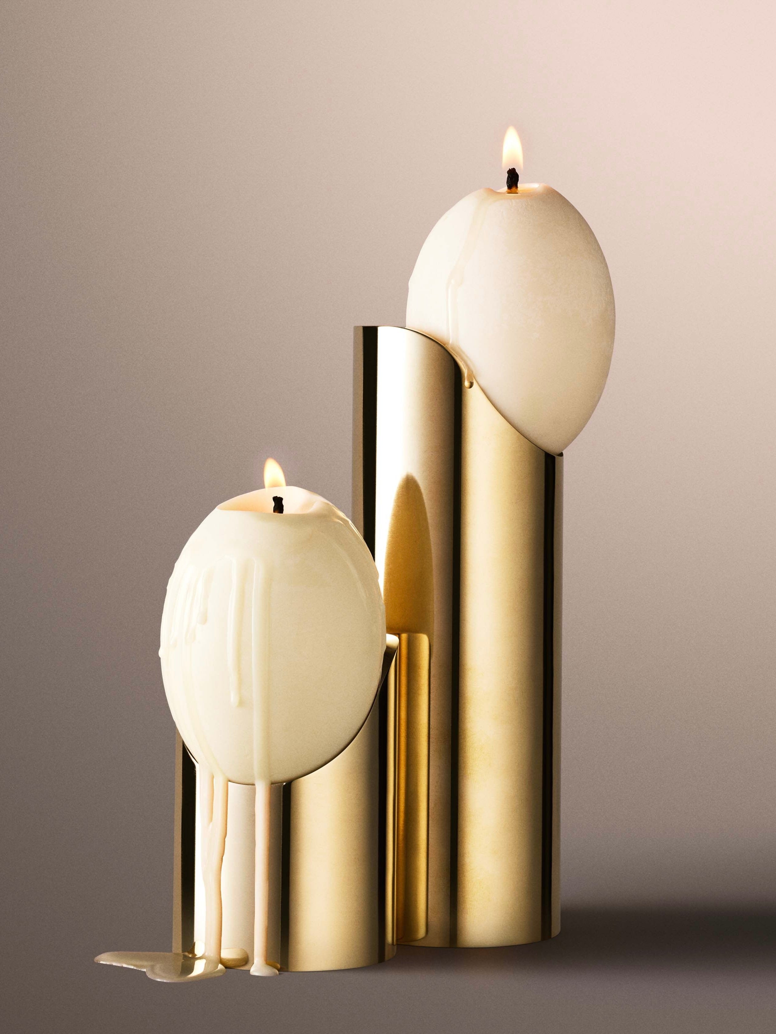 Two spherical white candles are alight, placed on modern, gold High Perch Holders by TĂȘte Studio of varying heights against a neutral background. One candle has melted wax dripping down its side, adding a touch of texture to the clean, elegant setup reminiscent of Noguchi sculptures.