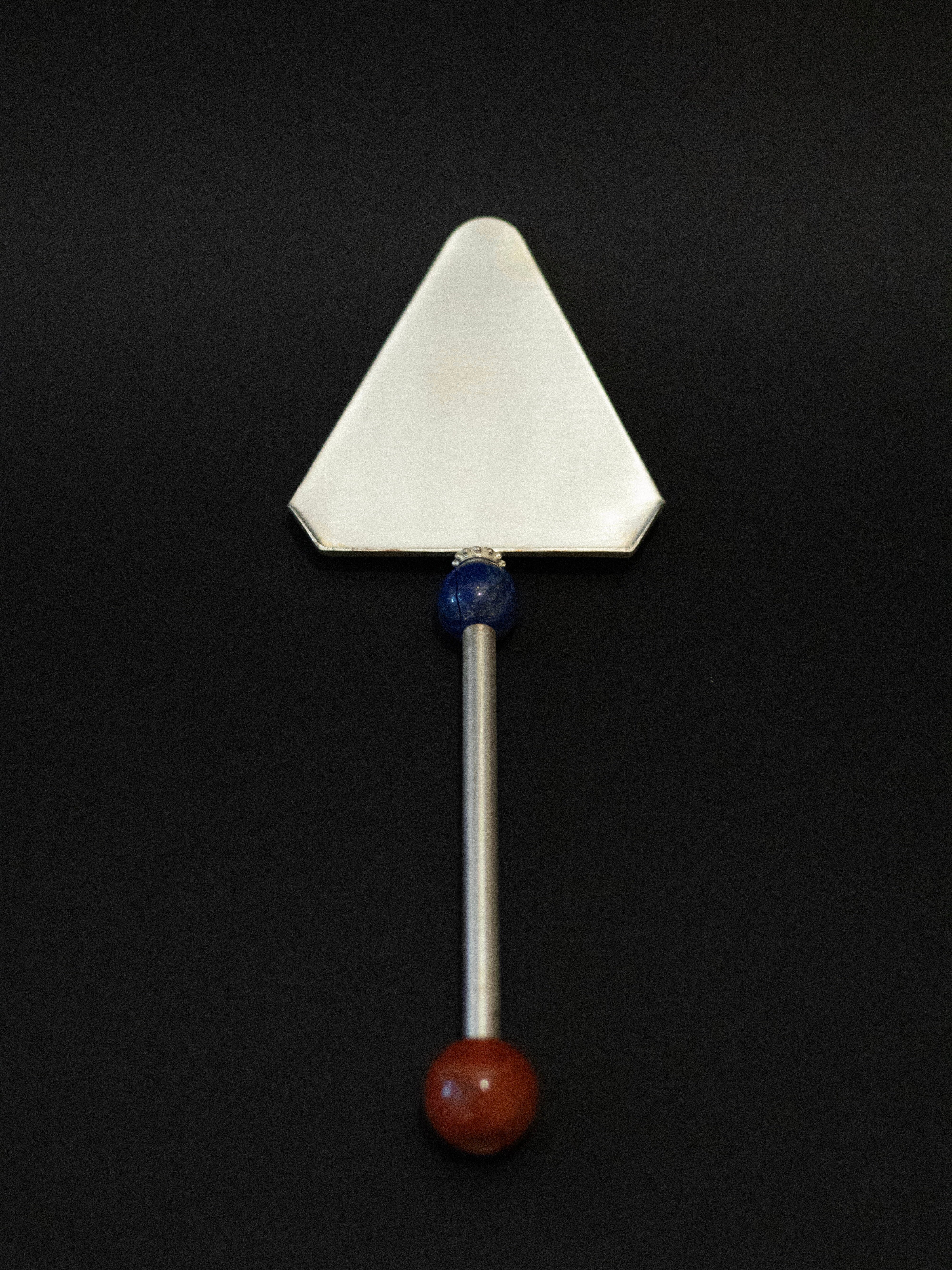 The Natalia Criado Cake Server features a stainless steel triangular blade, a straight handle with blue and red beads, and is uniquely displayed on a striking black background.