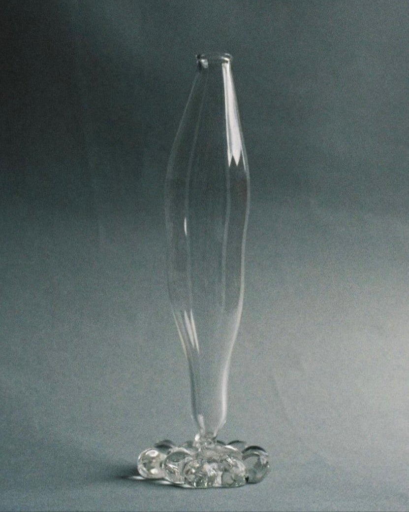 A tall, elegant Mouthblown Vase by Justine Menard stands on a flat surface, showcasing its hand-blown glass craftsmanship. The vase features a narrow, undulating body and an artistically shaped base with multiple rounded elements, giving it a unique and delicate appearance. The neutral gray background highlights the vase's transparency and long neck design beautifully.