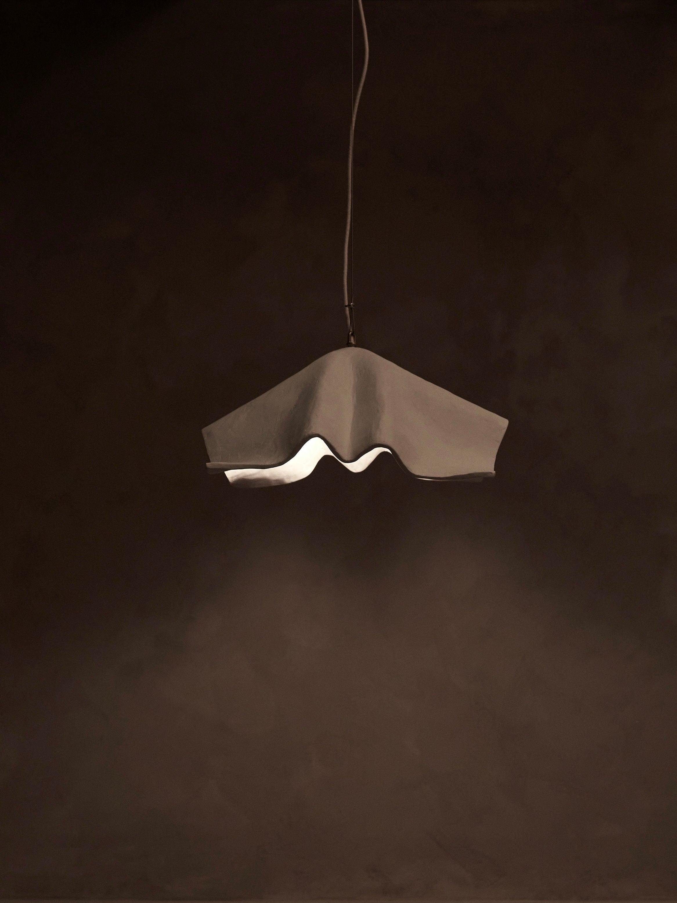The 'La Fleur' Pendant Light by Studio Tristan Kallas features a modern, wavy asymmetrical design against a dark backdrop. It emits a soft, warm glow with dimmable lighting, highlighting its unique shape and creating a serene ambiance for perfect mood control.