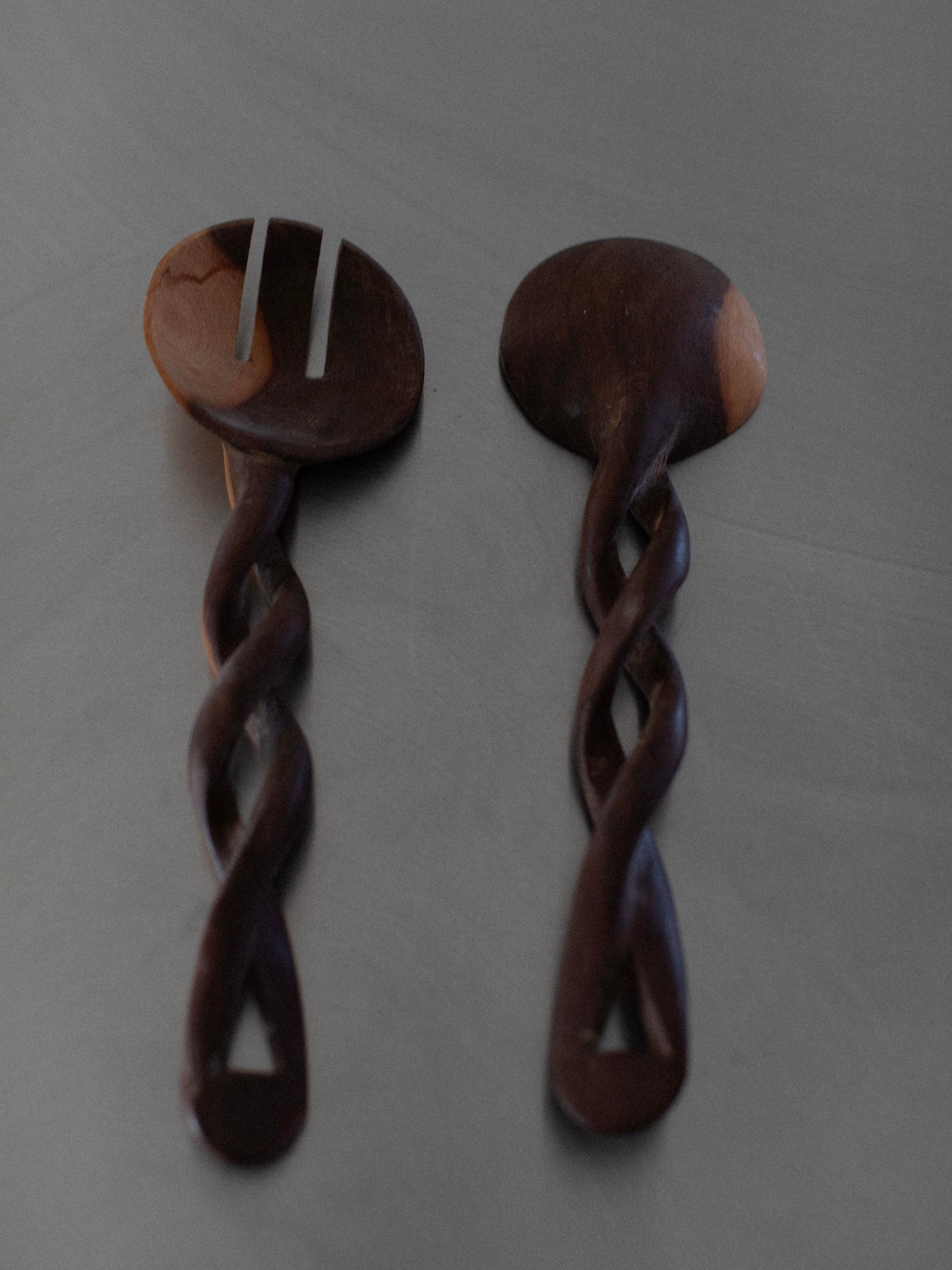 Two handcrafted Twisted Serving Spoons by Out For Lunch with intricate twisted handles rest on a flat surface. One is a serving fork with two prongs, and the other is a serving spoon. The wood appears dark with some lighter patches, showcasing its natural grain and subtle variations from the hand-carving process.