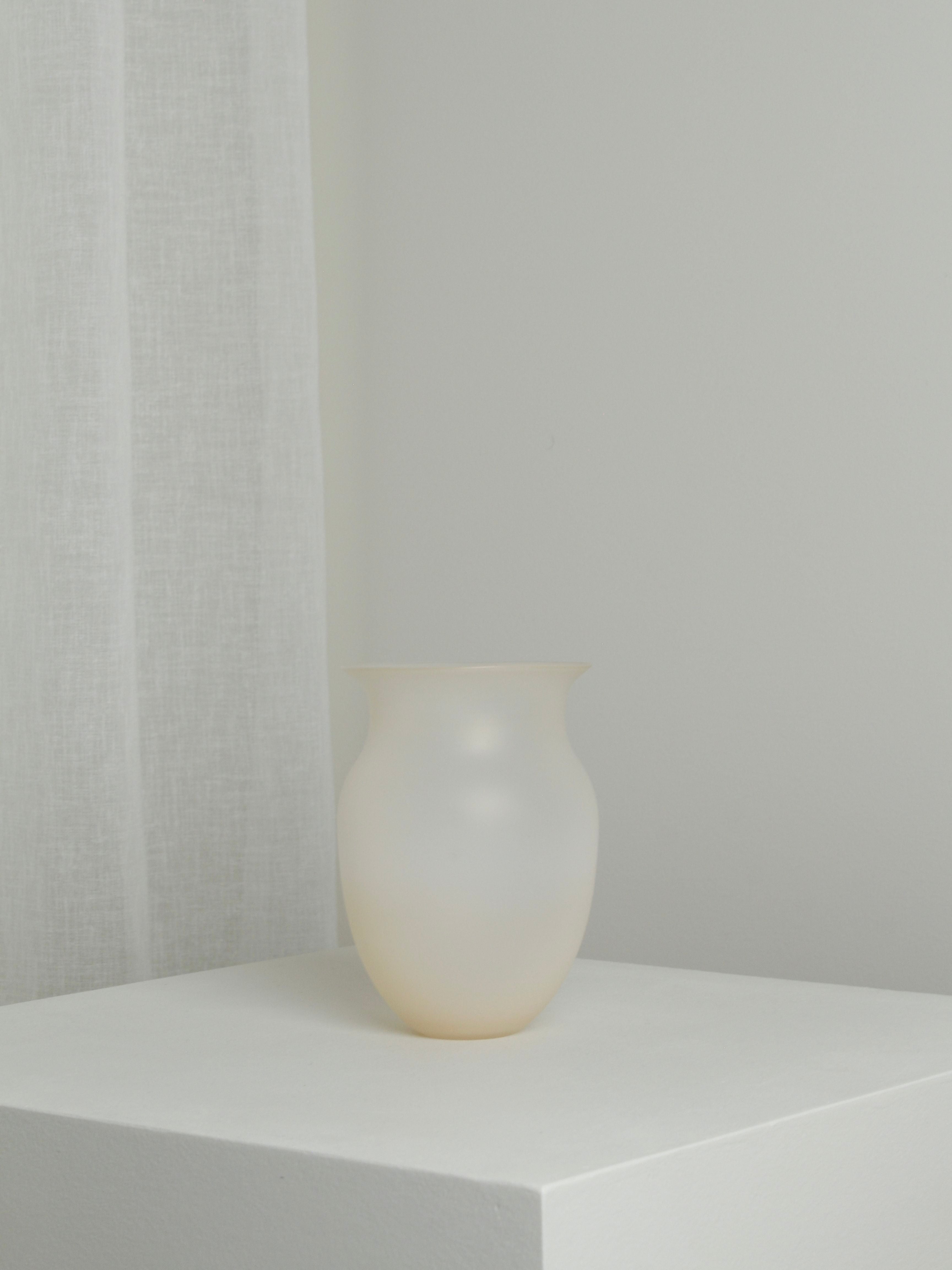 A Medium Vase in Sahara from Verre d'Onge, mouth-blown and translucent white, sits on a white pedestal in a minimalist setting. A white sheer curtain is visible in the background to the left. The overall color scheme is soft and neutral, emphasizing simplicity and elegance in this Sahara-inspired scene.