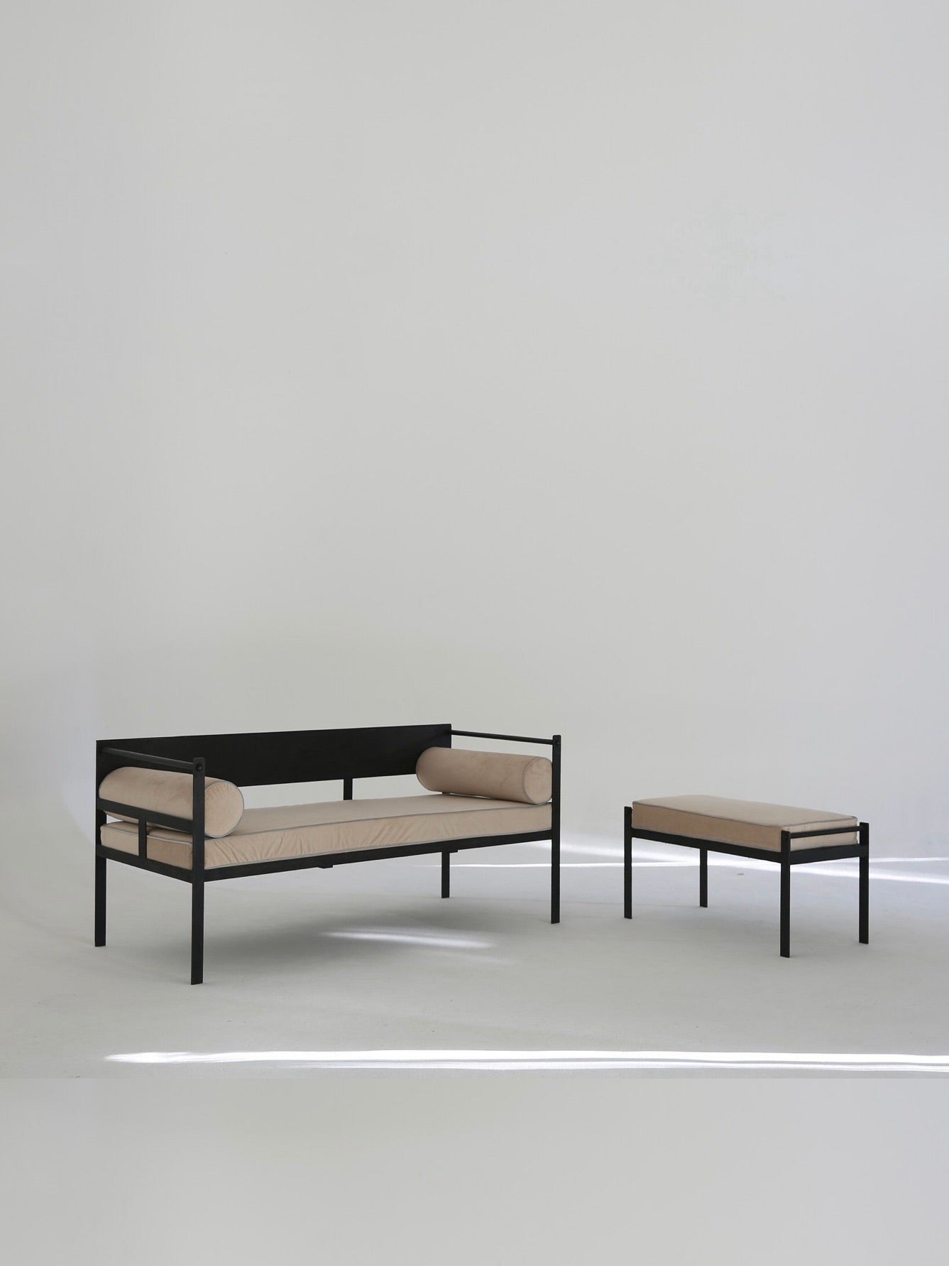 A minimalist room features the Ancien et Jolie Modernist Lounge Set with a Mathieu Mategot-inspired, black metal-framed daybed with beige cushions, two bolster pillows, and a matching rectangular ottoman. Simple light walls and floors enhance its minimalist design.