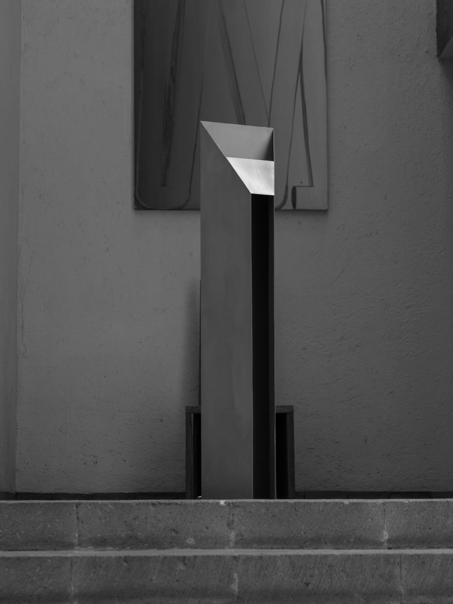 A tall, geometric sculpture with sharp angles stands on a stone step against a plain, light-colored wall. Above it hangs a rectangular artwork with abstract lines, reflecting minimalist artistry akin to Siete Studio's 'Fragua' Floor Lamp in its handcrafted elegance and modernity.