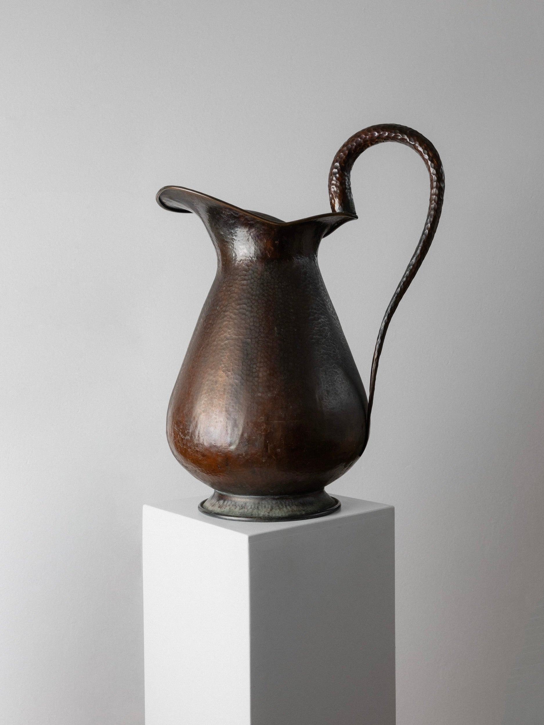 A tall, elegant piece from Bottega Jacobs—a Copper Handmade Jug/Vase from 1950s Italy—stands on a white pedestal against a gray background. Its curved spout and handle with a textured surface evoke vintage charm, adding an antique flair to the scene.