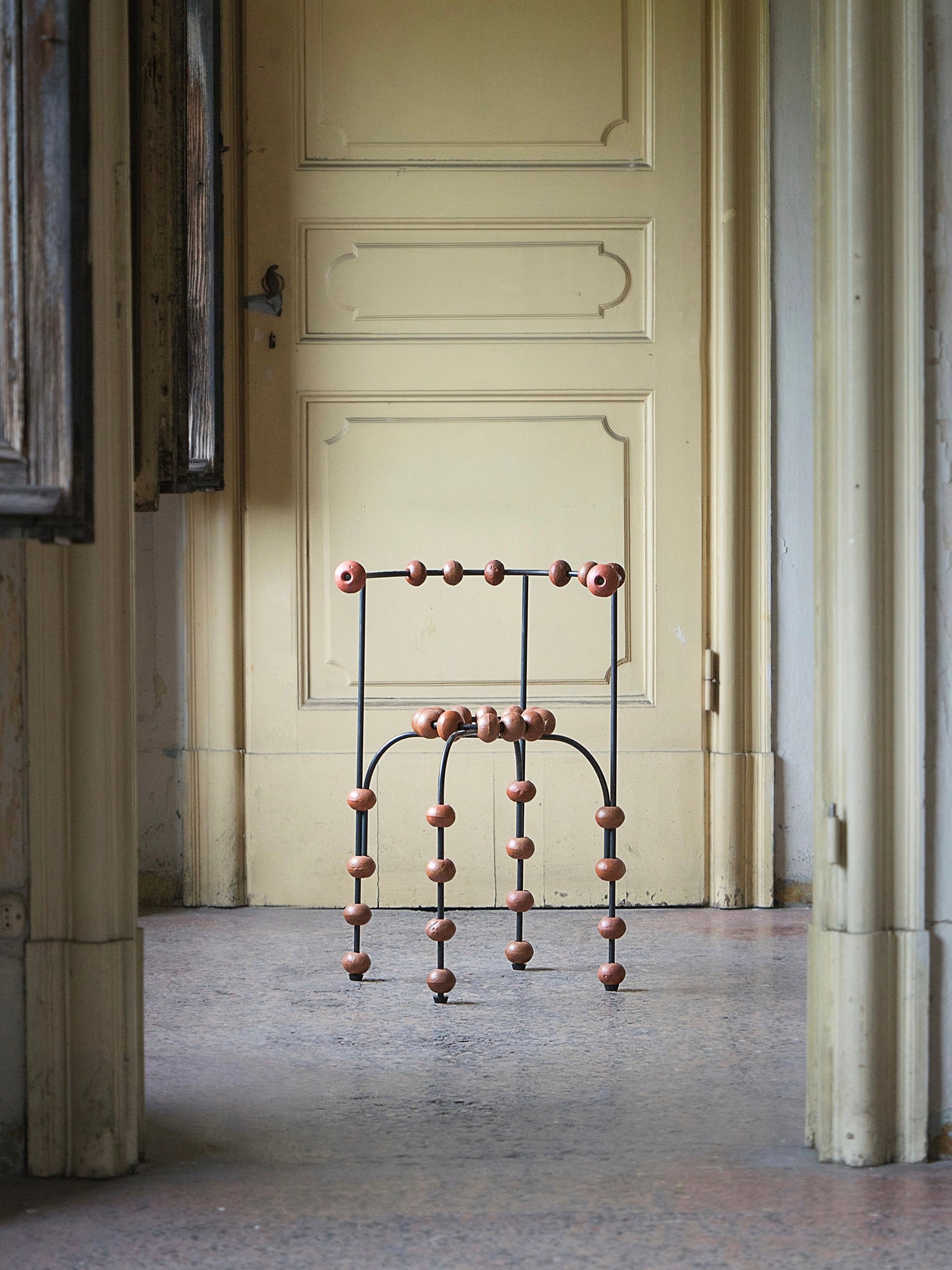 The "Buoys" Chair by Touch With Eyes stands in a dim hallway, featuring a metal frame with round wooden accents. A closed cream door and worn walls in the background enhance its vintage ambiance.