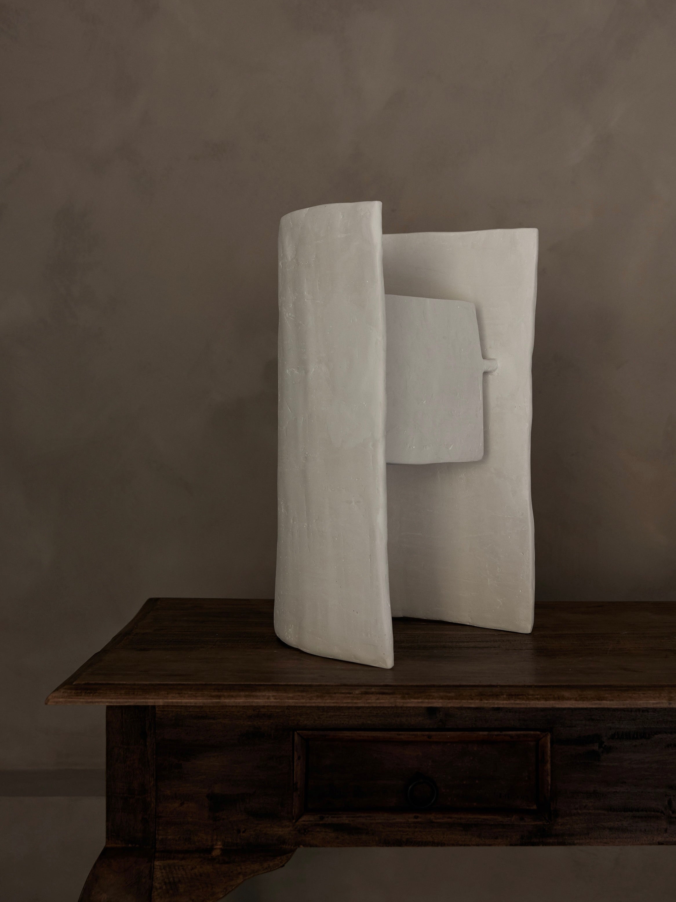 The Studio Tristan Kallas Crescent Table Lamp, with its minimalist curved and angular white design, rests on an antique wooden table. Indirect lighting accentuates its smooth, abstract form against a textured gray backdrop, beautifully contrasting with the rustic table surface.