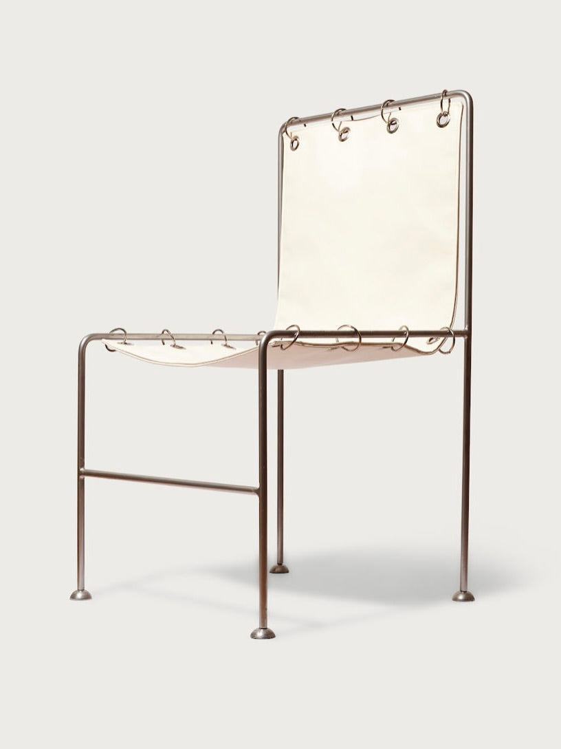 The Institution Chair by Panorammma features a minimalist design with a thin metal frame and beige fabric seat, enhanced by chromed steel rings and exposed loops. Its straight, slender legs add a modern-industrial appeal with clinical aesthetics.