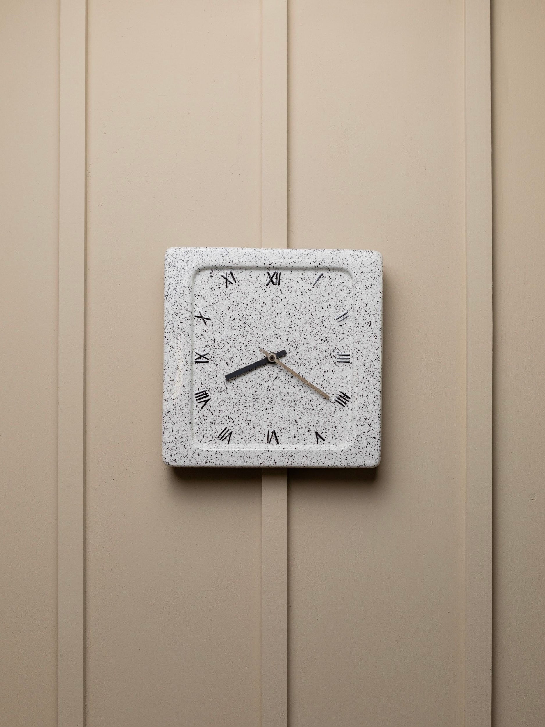 A Vintage Ceramic Wall Clock 1970s Italy by Bottega Jacobs with a speckled white frame hangs on a beige paneled wall, evoking timeless elegance. The clock face features Roman numerals and three black hands that tick away the hours in style.