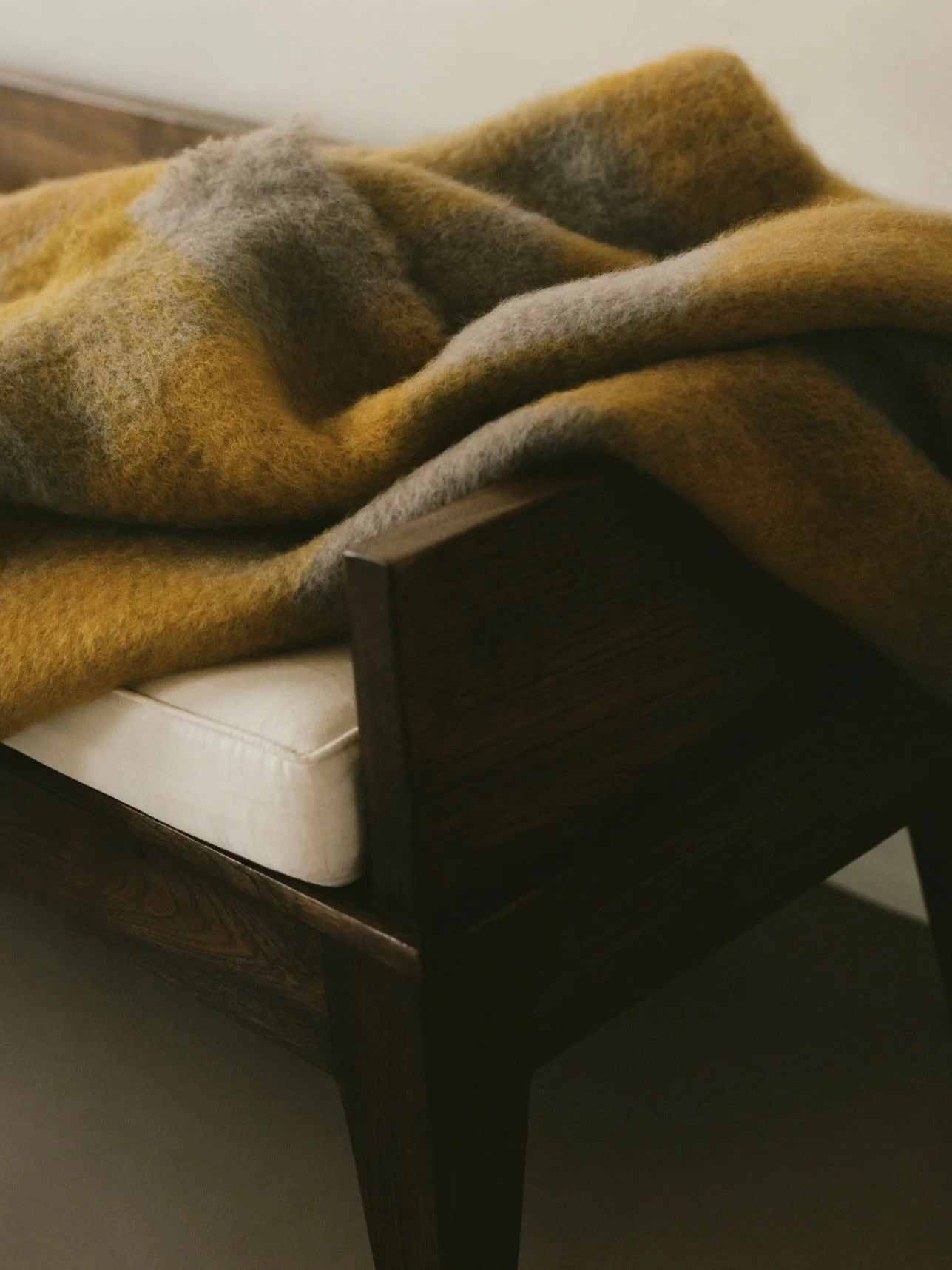 An "Arbor Fawn" Throw by The Wren in shades of yellow and gray is elegantly draped over the arm of a wooden sofa with a white cushion, creating an atmosphere that is both luxurious and inviting.