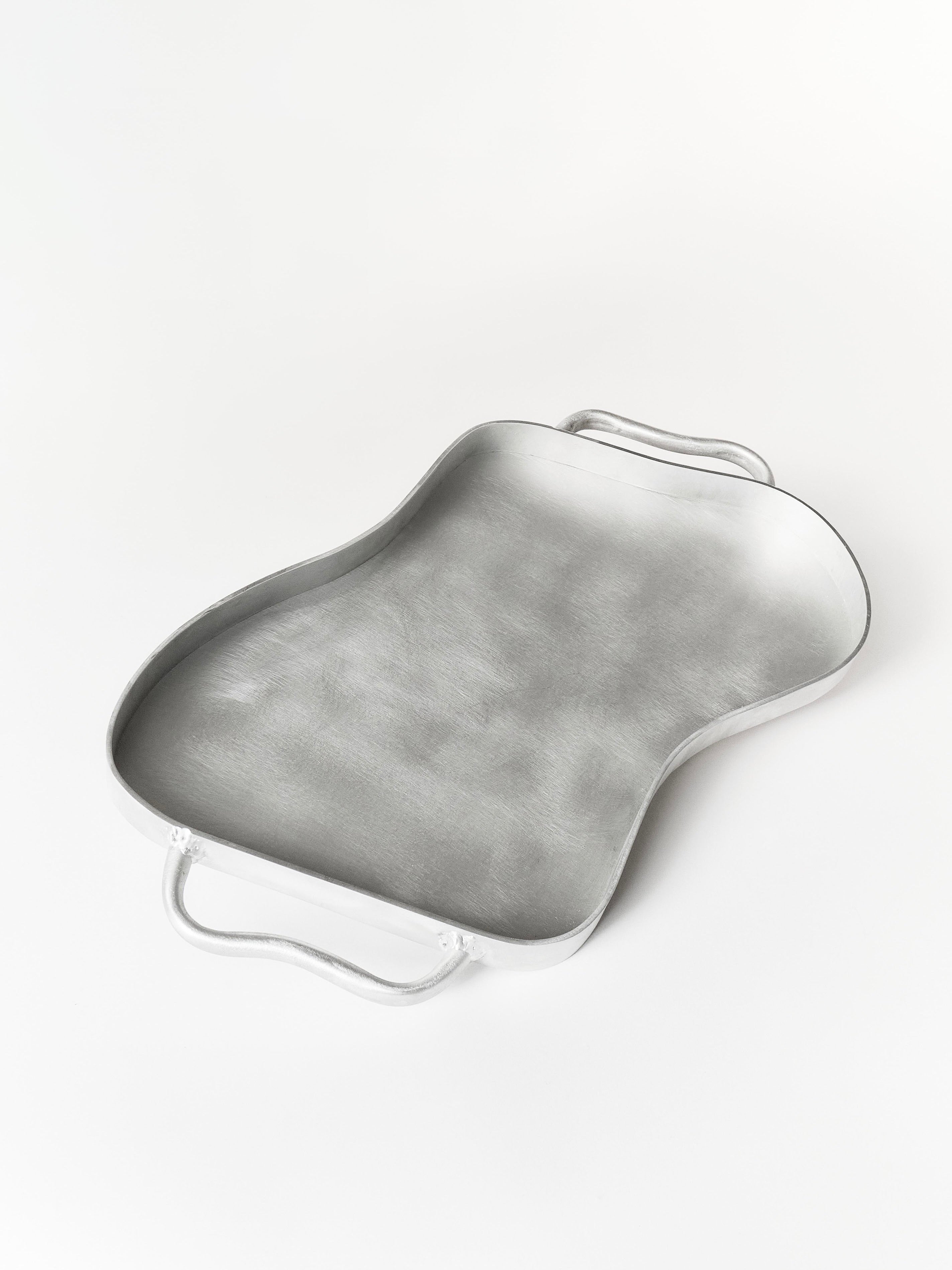 A metallic Serving Tray by Six Dots Design, with curved edges, two handles, an abstract irregular shape, and a smooth brushed texture, handmade in a North London workshop, rests on a plain white surface.