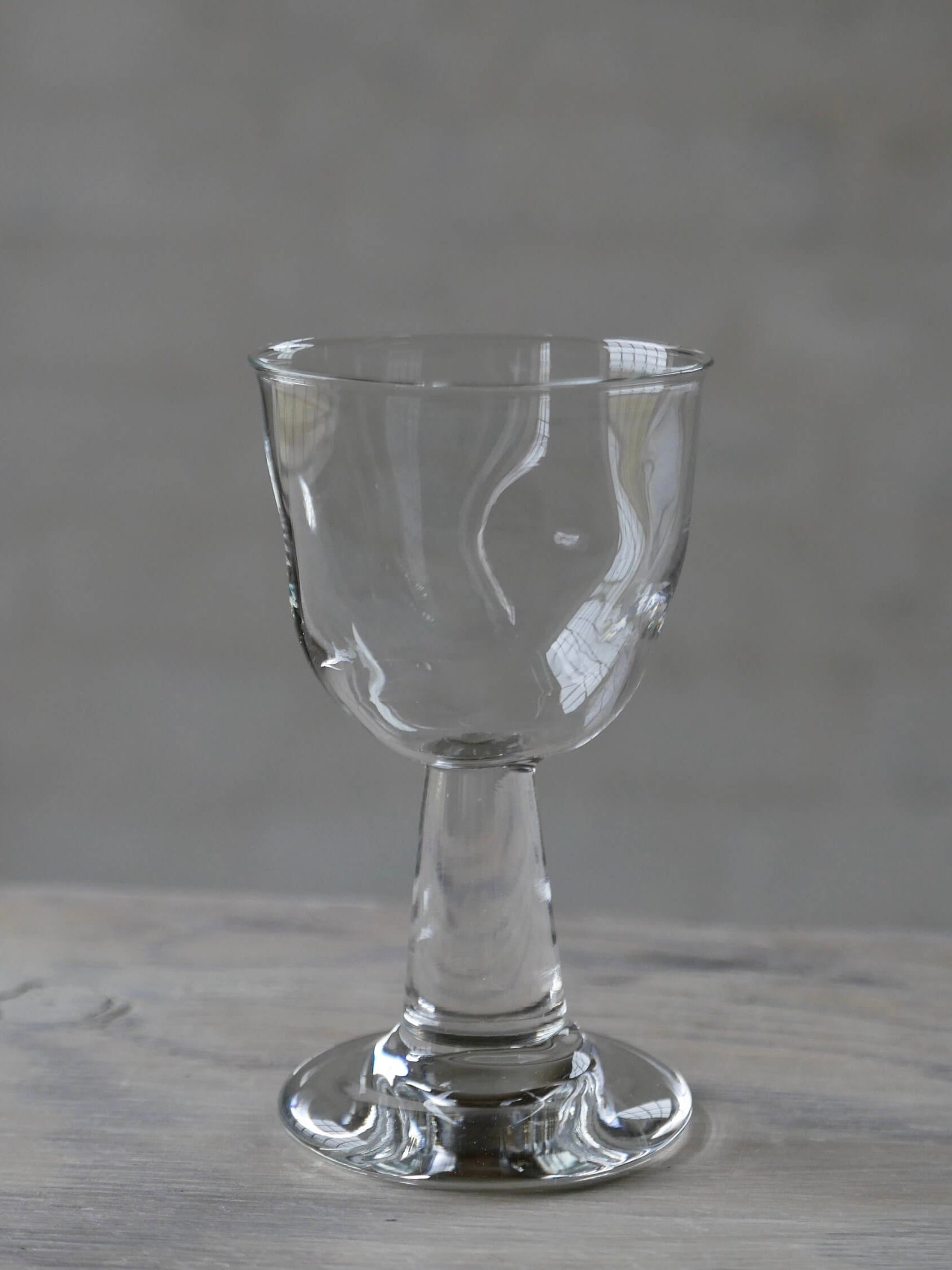 A clear, empty Silje Lindrup liqueur glass from the Liqueur Glass - Set of 4, featuring a short stem and wide base, stands on a wooden surface against a blurred gray background, promising an exquisite drinking experience.