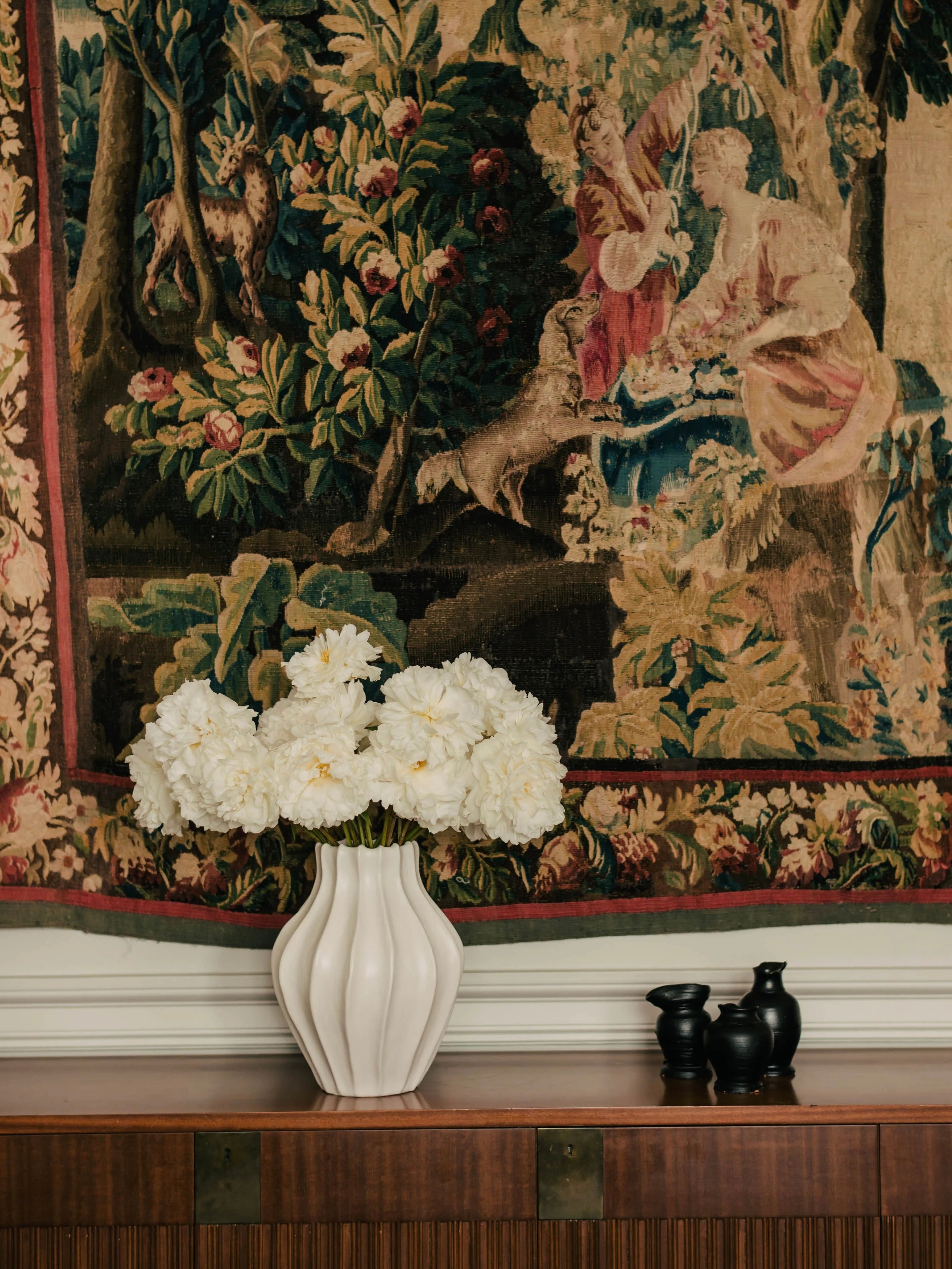 A Ransom & Dunn Medium Cloak Vase filled with white peonies sits gracefully on a wooden cabinet. Behind it, an intricately detailed tapestry showcases figures and lush greenery, while two small black decorative objects complete the scene, seamlessly blending into a tasteful ceramic collection.