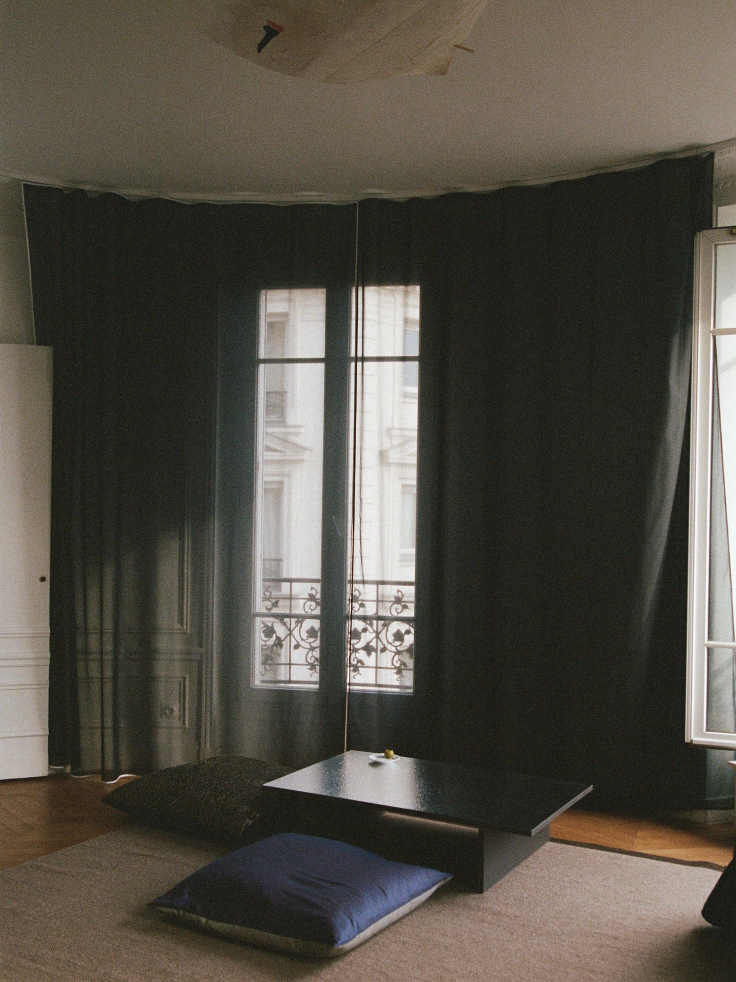 In a minimalist room with large dark curtains partially open to reveal a balcony and window, a low Mara Sister Coffee Table – Silver by TEGET sits on a beige rug surrounded by dark cushions. Light streams in, crafting a cozy and serene atmosphere.