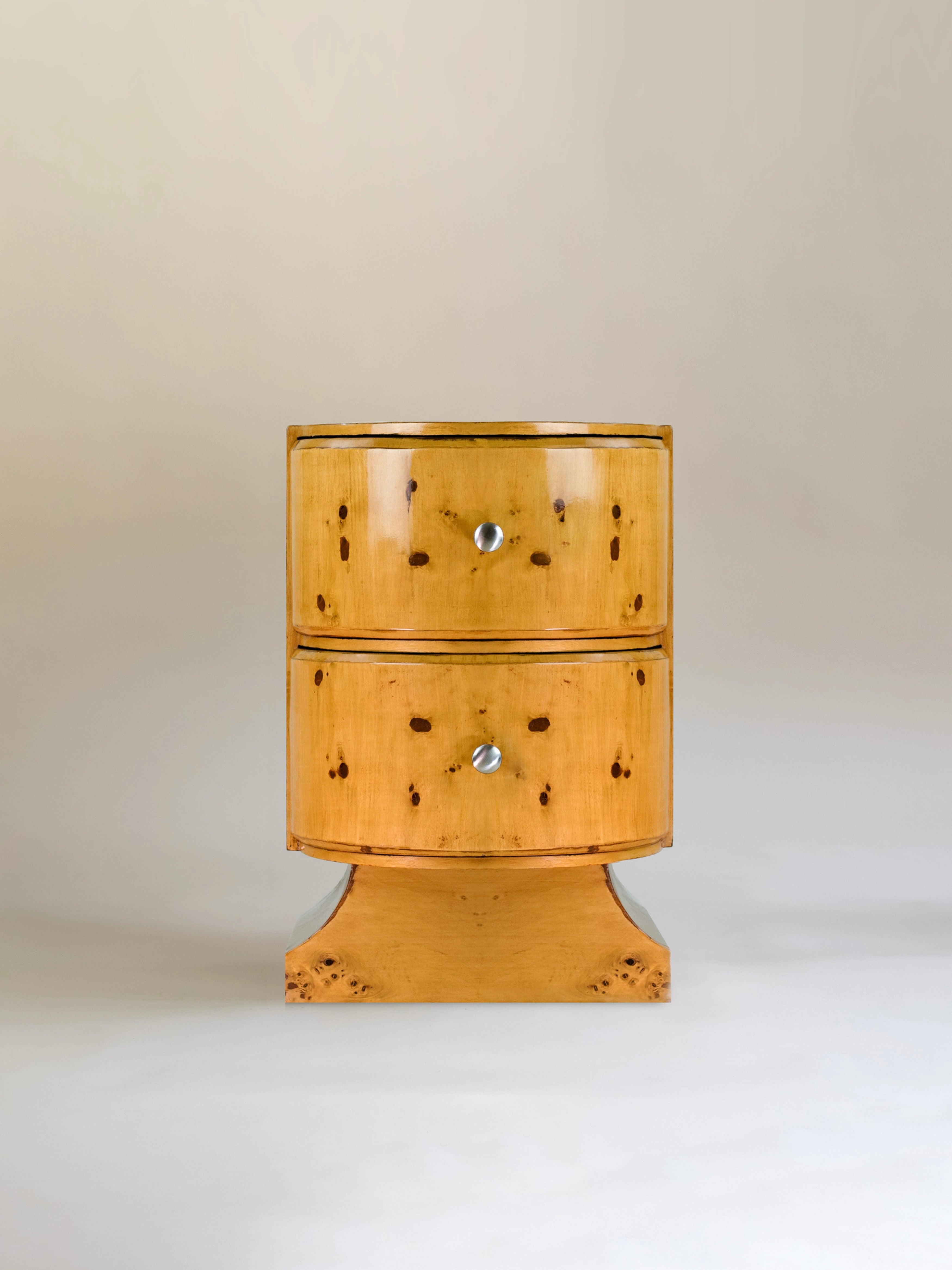 Meola Interiors' Burl Drawer is an Art Deco-style wooden chest featuring two drawers with round silver knobs, a warm glossy finish that highlights the burl wood veneer, and distinct natural patterns. Its curved, symmetrical base exudes a minimalist aesthetic and vintage appeal.