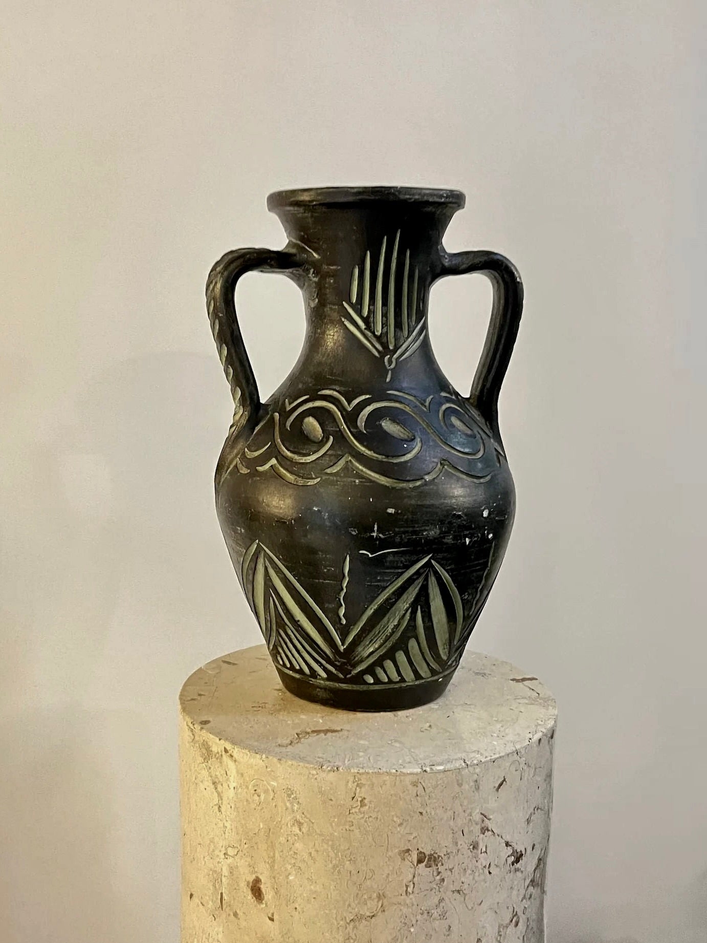 A rare black ceramic "European Large Carved Vase" from Aesthete Label, featuring two handles and intricate beige designs, including zigzags and wave patterns, is displayed on a rough, cylindrical stone pedestal. The minimalistic background accentuates the vase's detailed craftsmanship.