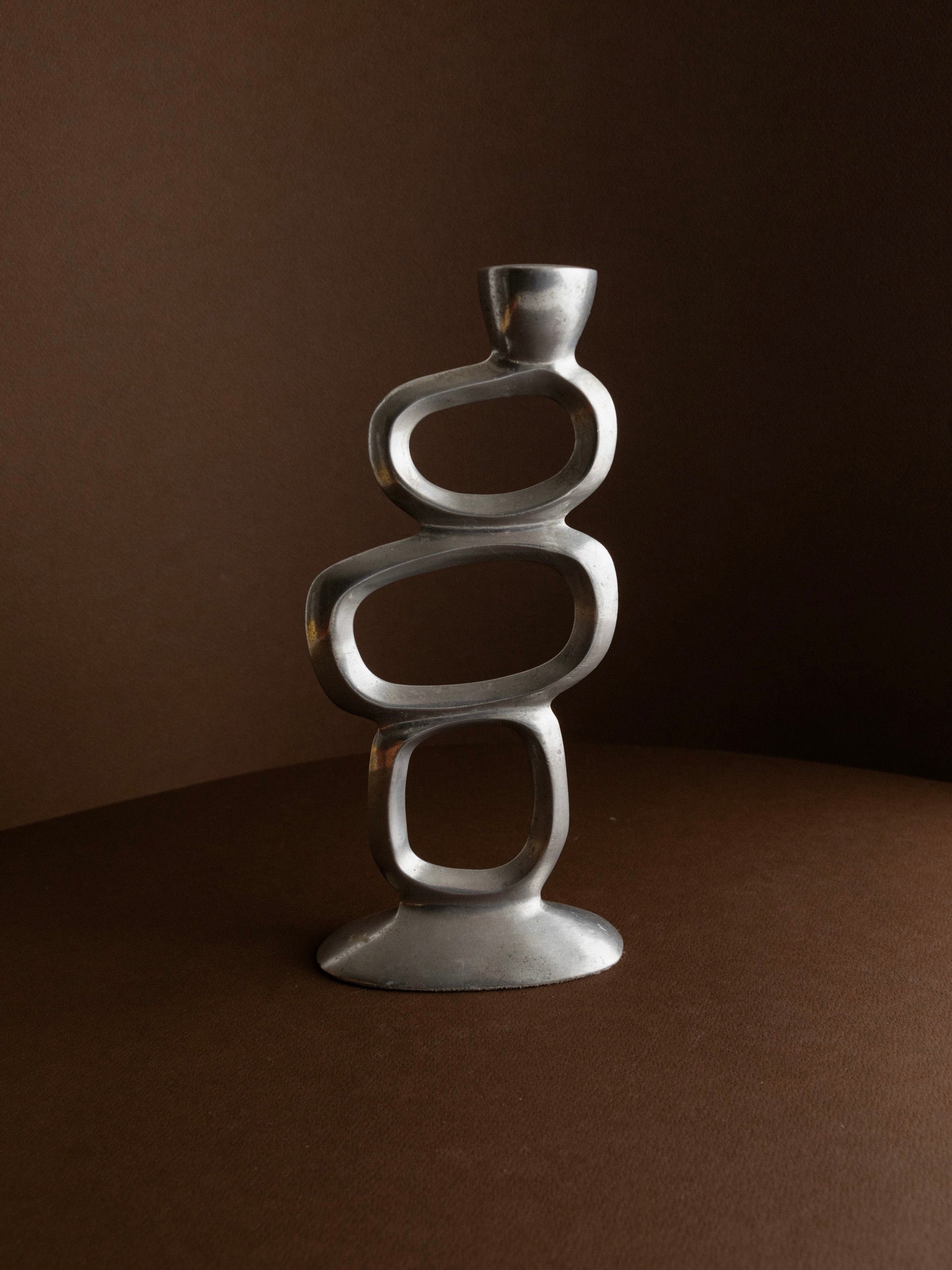 The Matthew Hilton Candleholder 70s by Bottega Jacobs is a modern, sculptural metal piece with an abstract, interconnected loop design. It evokes a mid-century vibe, placed on a smooth brown surface against a dark brown background.