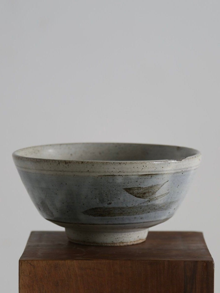 A Leach Pottery Bowl by Ancien et Jolie rests on a wooden block, showcasing a wide rim and speckled gray glaze. It features subtle avian motifs and charming imperfections for an artisanal look reminiscent of British 1960s designs, set against a plain, softly lit background.