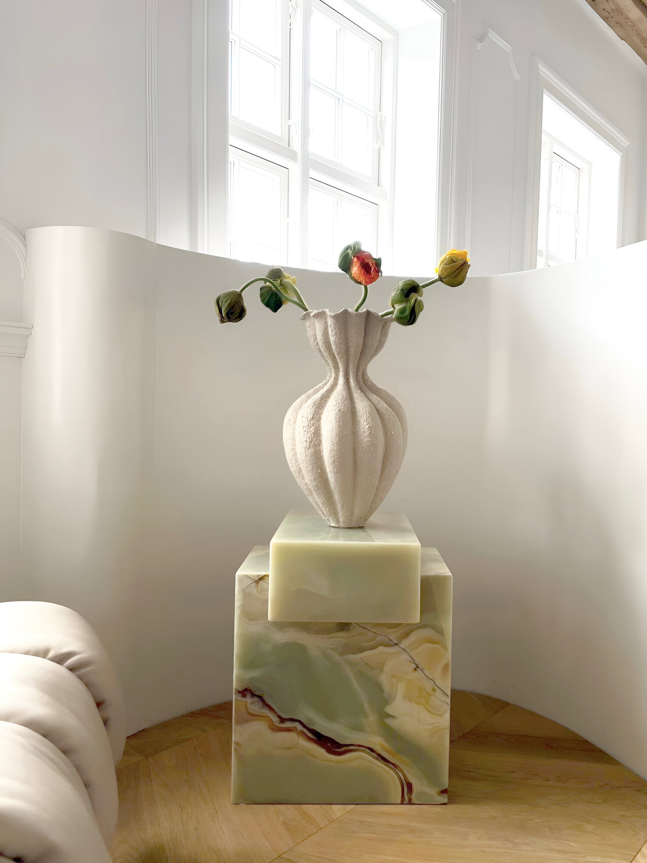 A sculpted cream-colored vase with drooping tulips sits on the Gaia Green Onyx Marble Coffee Table by Marbera in a bright, minimalist room with curved white walls and large windows, embodying modern decor.