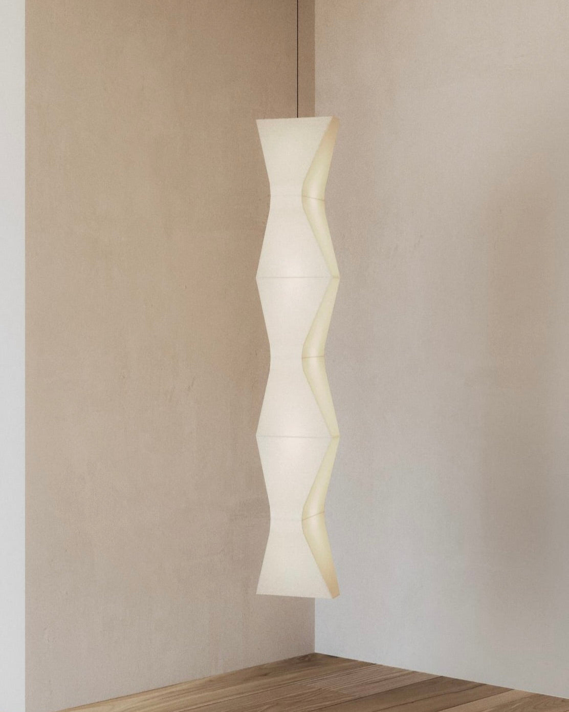 An "Arête" Pendant Vertical Line by Asca Studio with a geometric design enhances a minimalistic room corner. Its wheat-colored linen shade gently glows, complementing the light wood flooring and plain beige walls, creating a warm and inviting atmosphere.
