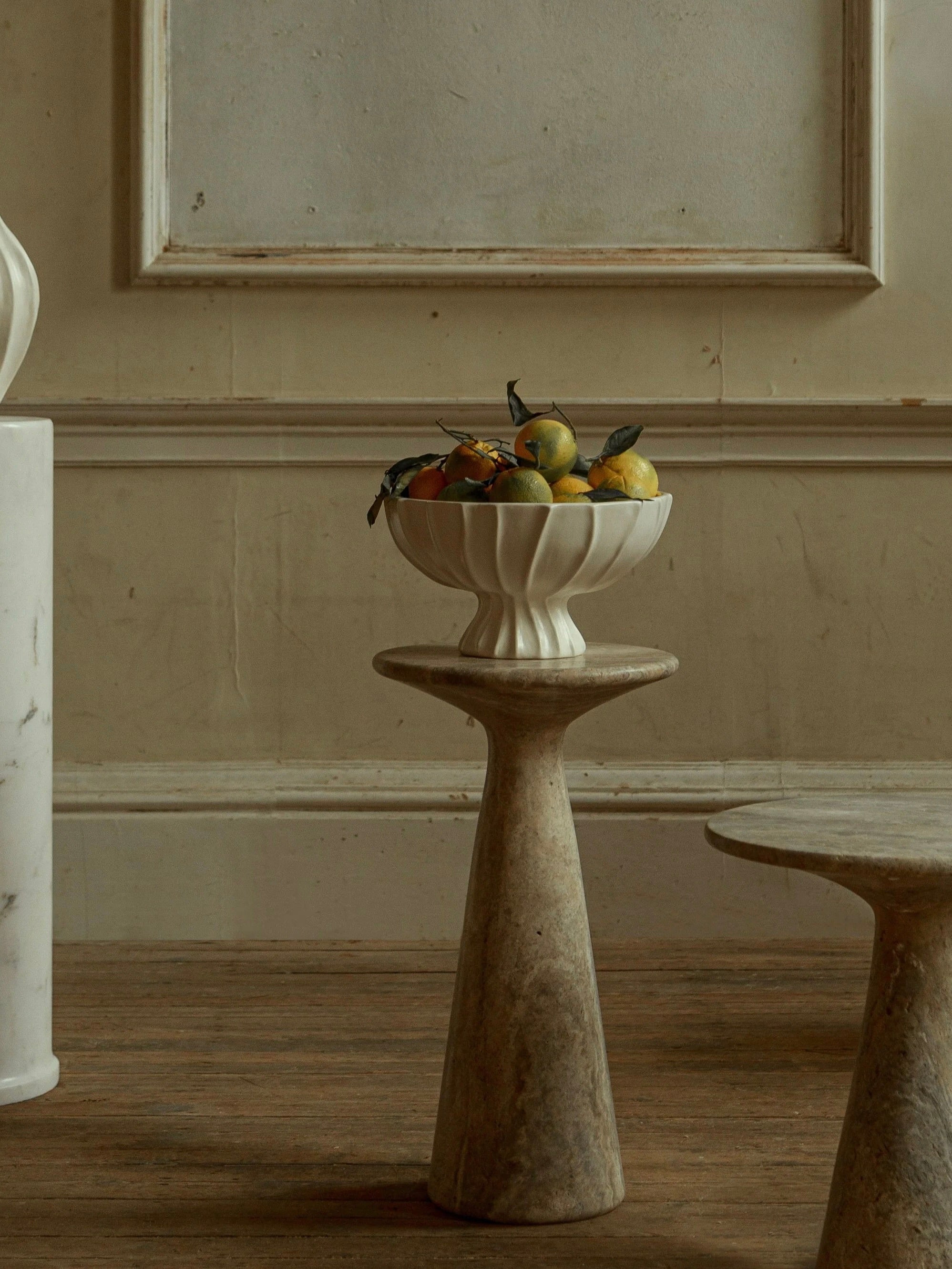 A decorative fruit bowl holding yellow and red fruits sits on the Juno Tall Travertine Side Table by Ransom & Dunn, accompanied by a matching stool. The setup, handcrafted in Europe, is set against a plain beige textured wall, creating an elegant and minimalistic atmosphere.