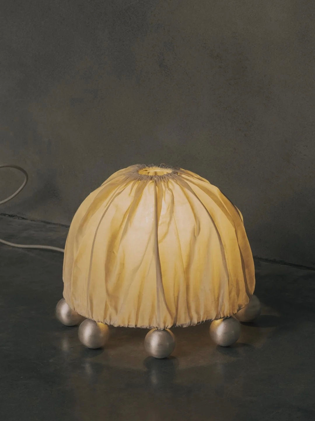 The "ALTER" Table Lamp by Kaytar features a fabric-covered dome emitting a warm glow, supported by spherical feet. With its sculptural design on a dark reflective surface against a muted background, it exudes an artistic charm reminiscent of alchemy symbols.