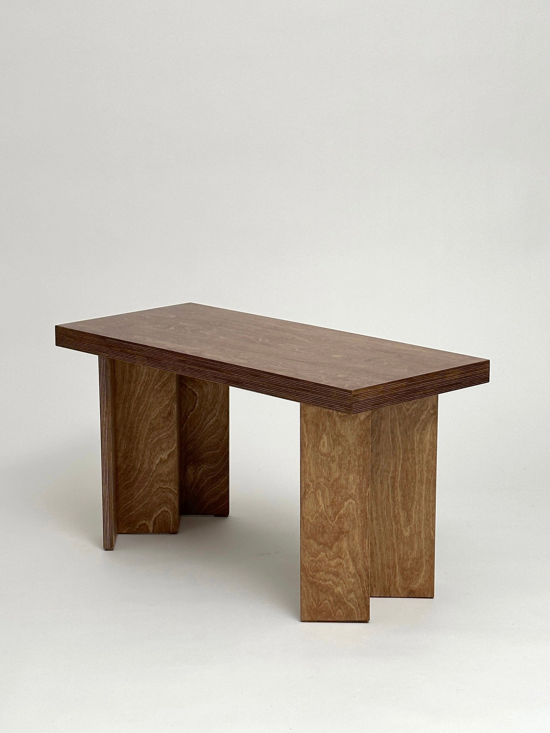 Introducing the Single Zig-Zag Bench from GOONS: a wooden bench with a smooth, rectangular top supported by two robust, geometric legs, crafted by a Paris-based design studio. Set against a neutral background, the wood's natural grain pattern imparts a rustic charm.