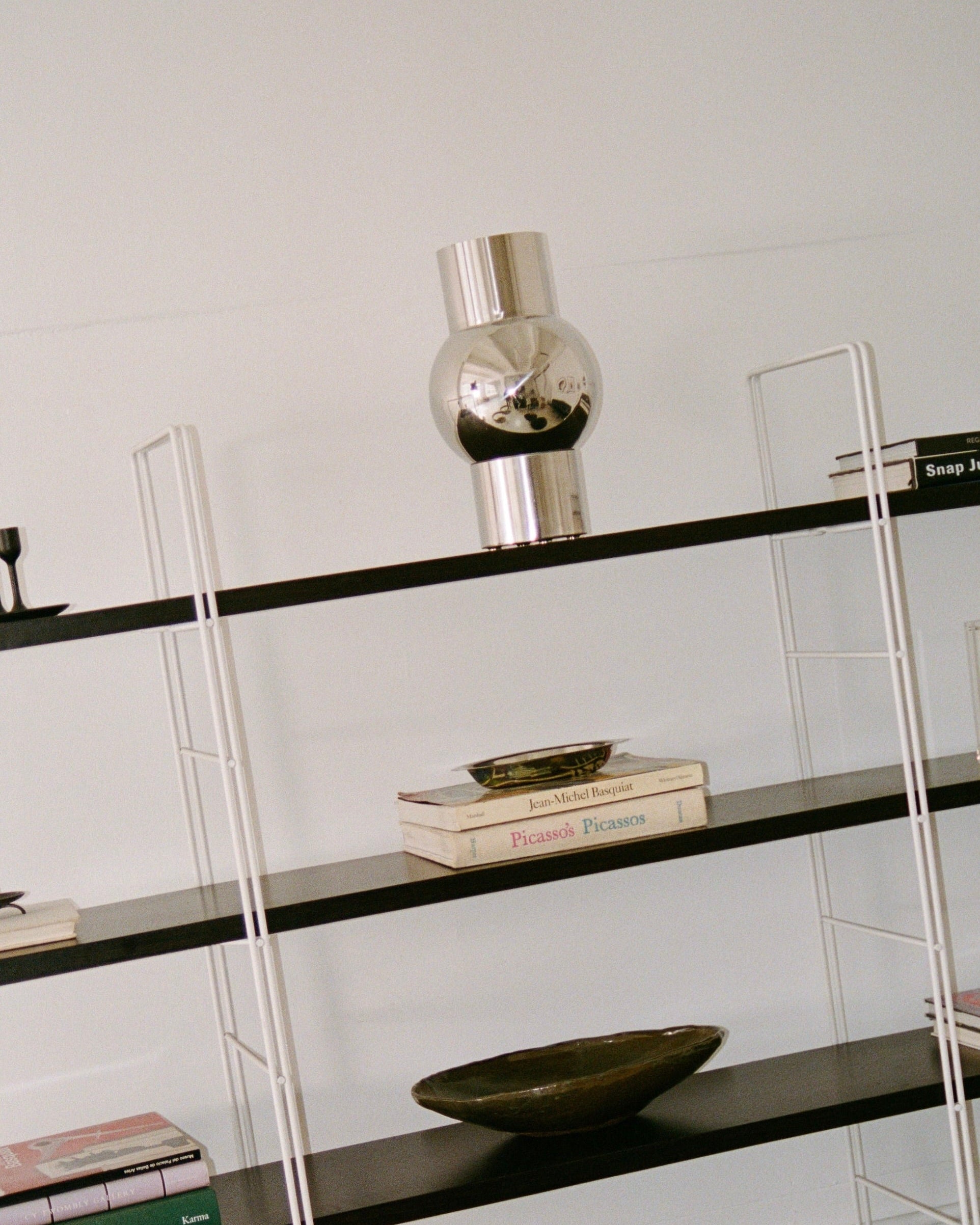 A modern black and white bookshelf with several items. The top shelf holds a reflective stainless steel decorative piece. The middle shelves have books, including "Picasso" and "Picassos," along with the MUHLY-brand Mica Vase in a greenish tone. The lower shelves feature more books, including one with a pink cover.