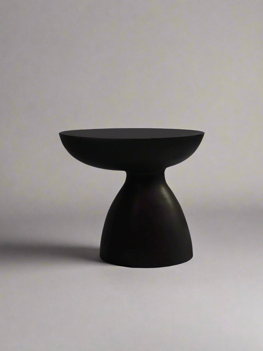The "Cleo" Ceramic Side Table by Ransom & Dunn is a minimalist black stool with a matte black finish, showcasing a rounded top and a conical base, set against a plain gray background.