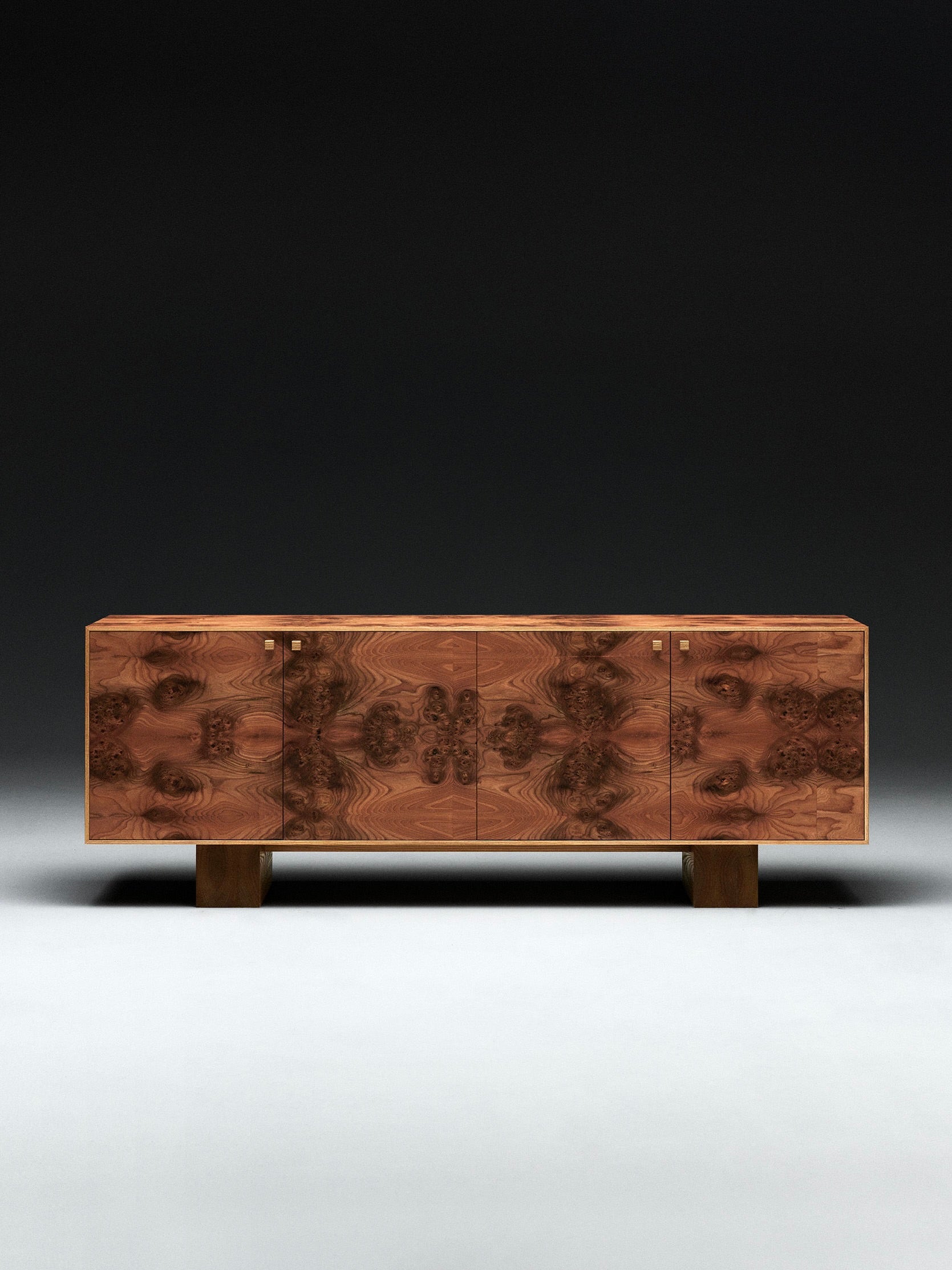 The 'Elm Burl' Sideboard by Axel Wannberg features a symmetrical, intricate wood grain pattern on its doors, four square legs, and small square handles, all set against a dark gradient background.