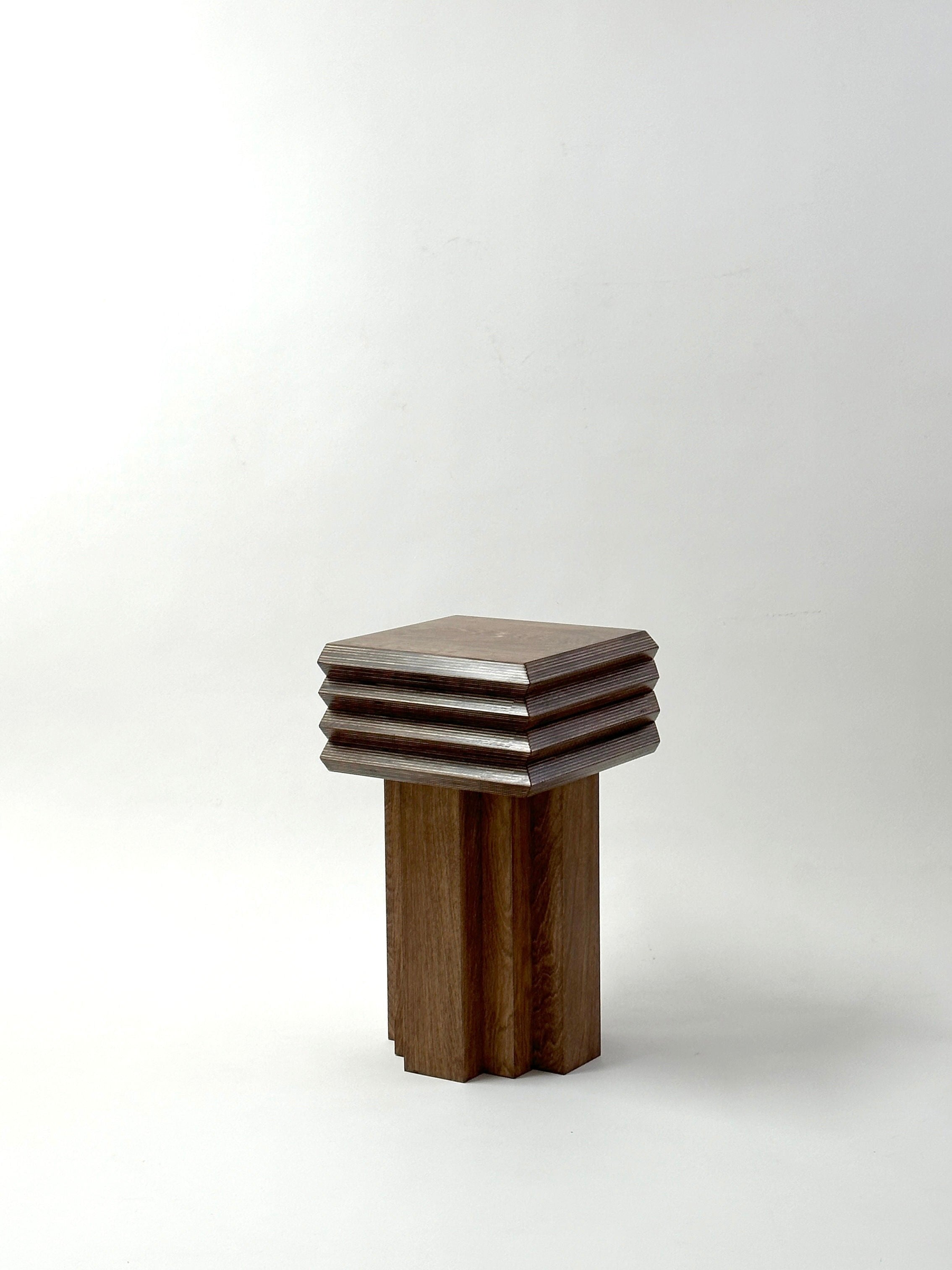 Presenting the MM Stool - Dark Oak by GOONS: a wooden stool with a geometric design. Constructed from dark oak, it showcases layered square shapes with beveled edges. Its rectangular supports form the base, enhancing its modern, minimalist look against a plain backdrop.