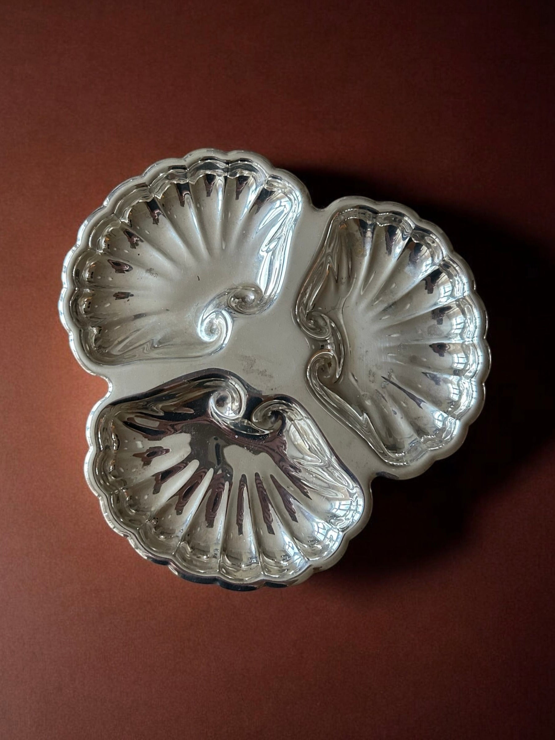 A Flock Studio Triple Shell Server, finished in silver and featuring three compartments, rests on a brown surface reflecting light elegantly.