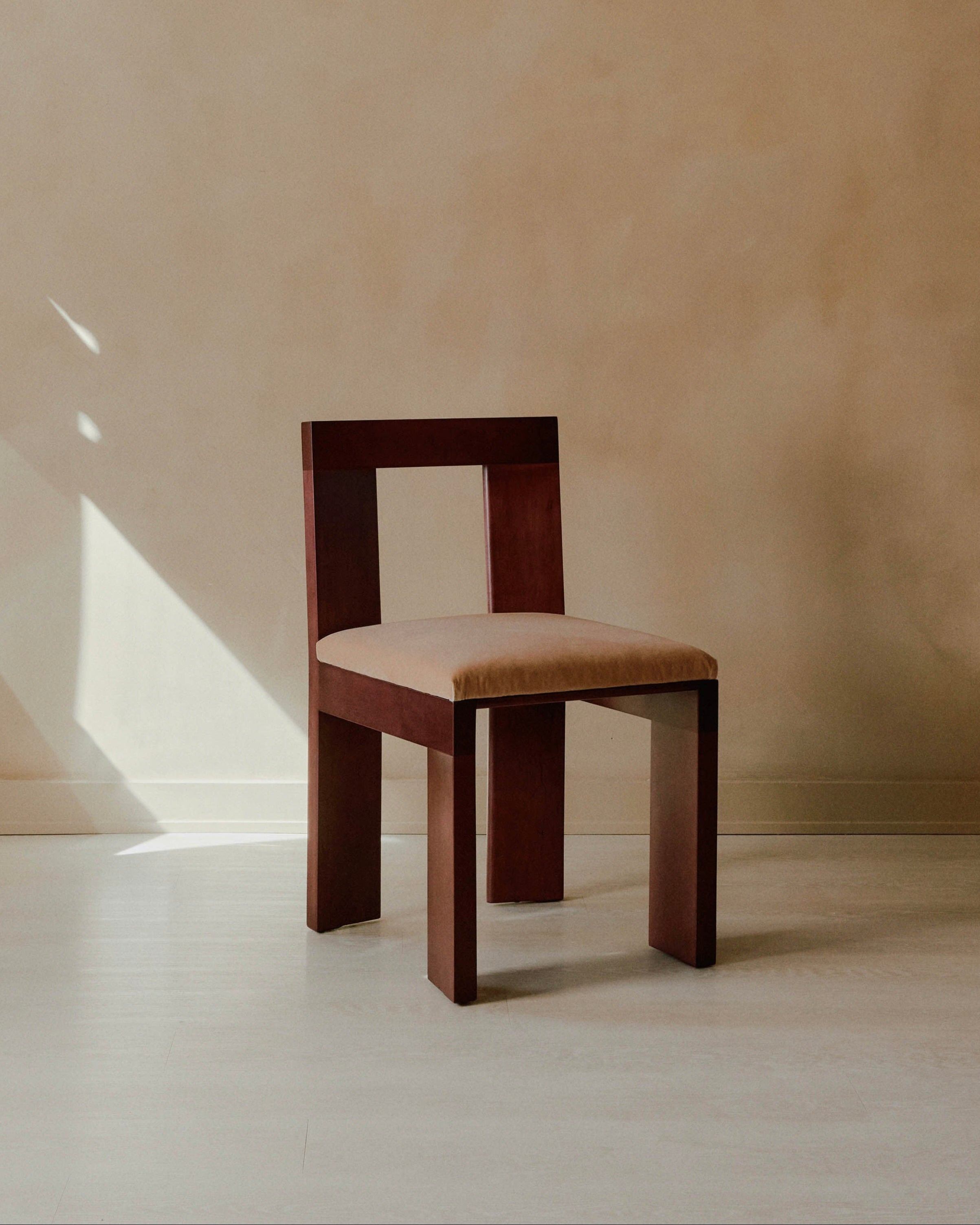 A simple Raja Chair by Juntos Projects, featuring a wooden plank design and a light beige upholstered seat, is positioned against a beige wall. It is illuminated by natural light streaming in from the left, casting soft shadows on the floor.