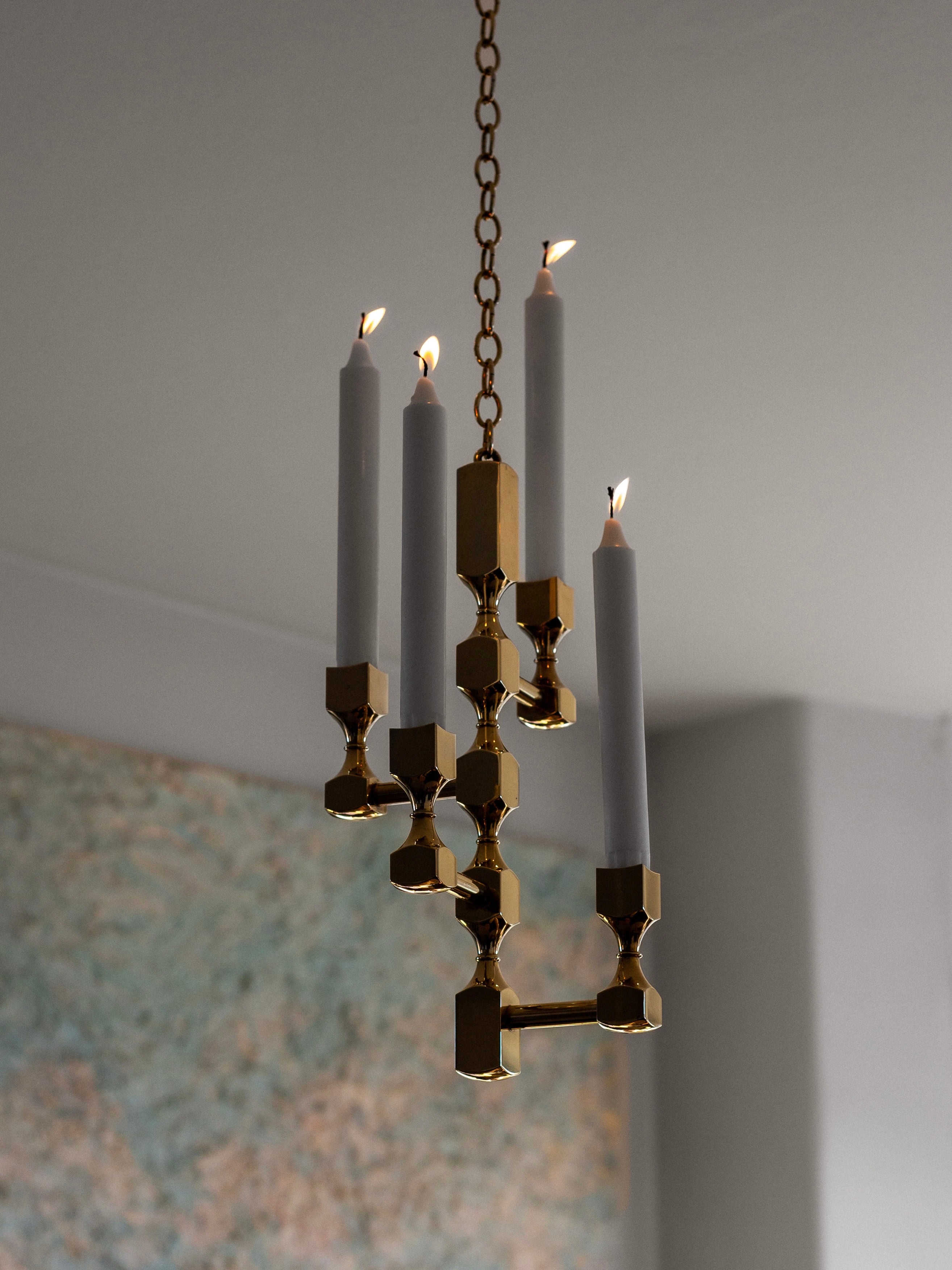 A Sculptural Brass Chandelier by Spigel, featuring a timeless design with hexagonal patterns, holds six candle-shaped lights. It hangs elegantly from a chain against a neutral-toned wall, highlighting the classic Bernt Karlsson aesthetic, with large blurred artwork subtly framing the scene.
