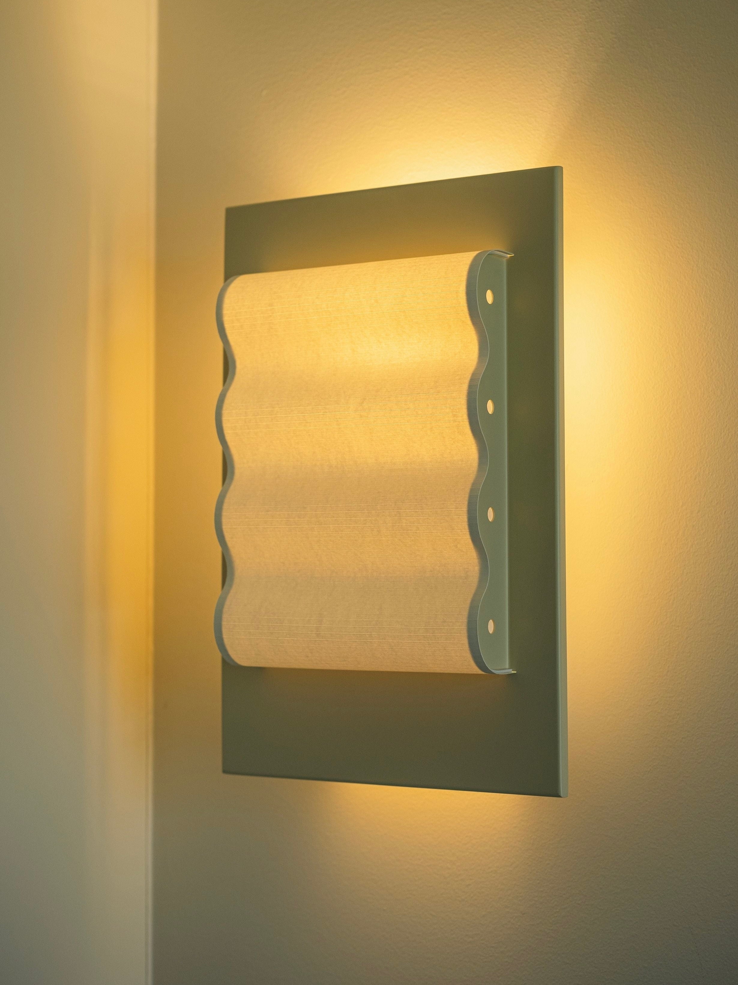 The Wall Light Frame OW M by Violaine d'Harcourt emits a warm, soft light. This modern light fixture features a rectangular backplate and a wave-patterned, frosted shade that diffuses the light evenly across the wall. Handcrafted with a minimalist design, this elegant wall sconce adds an elegant touch to any room.