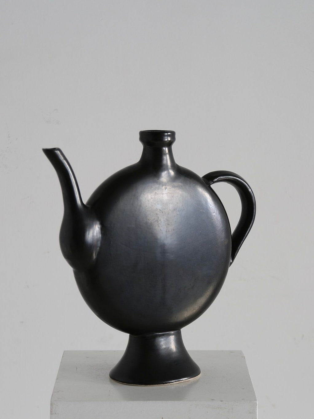 The Ancien et Jolie French Glazed Pitcher, with a minimalist matte black ceramic design reminiscent of 1960s France, features a round body, slender spout, and an elegant handle. It elegantly sits on a small white pedestal against a light gray backdrop.
