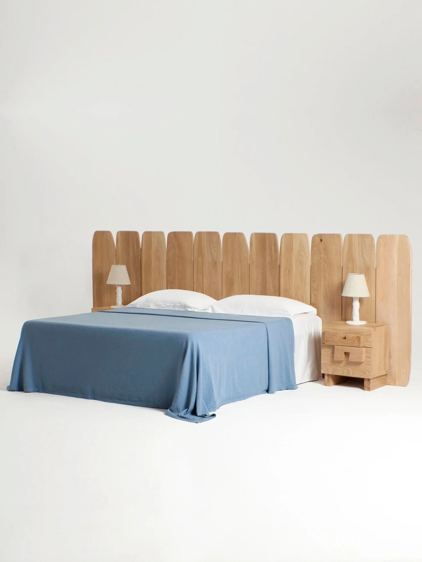A modern bedroom setup featuring a large wooden king-size bed with the Madeira Oak Headboard Large from Project 213A, handmade in Portugal. The bed is adorned with white sheets and a blue bedspread. Two wooden bedside tables with matching lamps are placed on each side of the bed against a plain white background.
