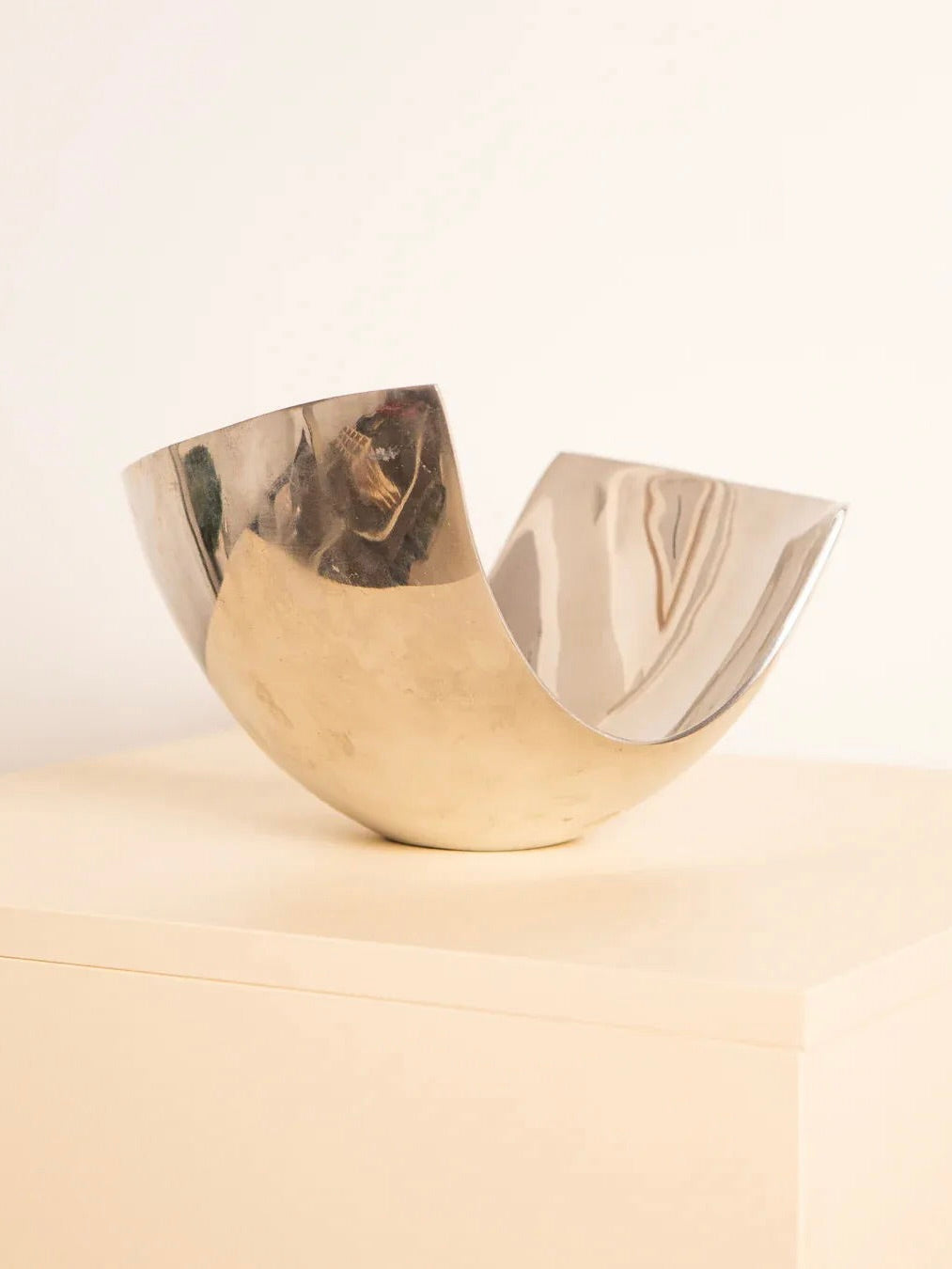 A "U"-Shaped Fruit Basket 80's, crafted by Treaptyque, features a gleaming metallic finish with a reflective surface. It rests elegantly on a light-toned rectangular stand against a simple backdrop, showcasing an asymmetrical, abstract form inspired by retro design aesthetics.