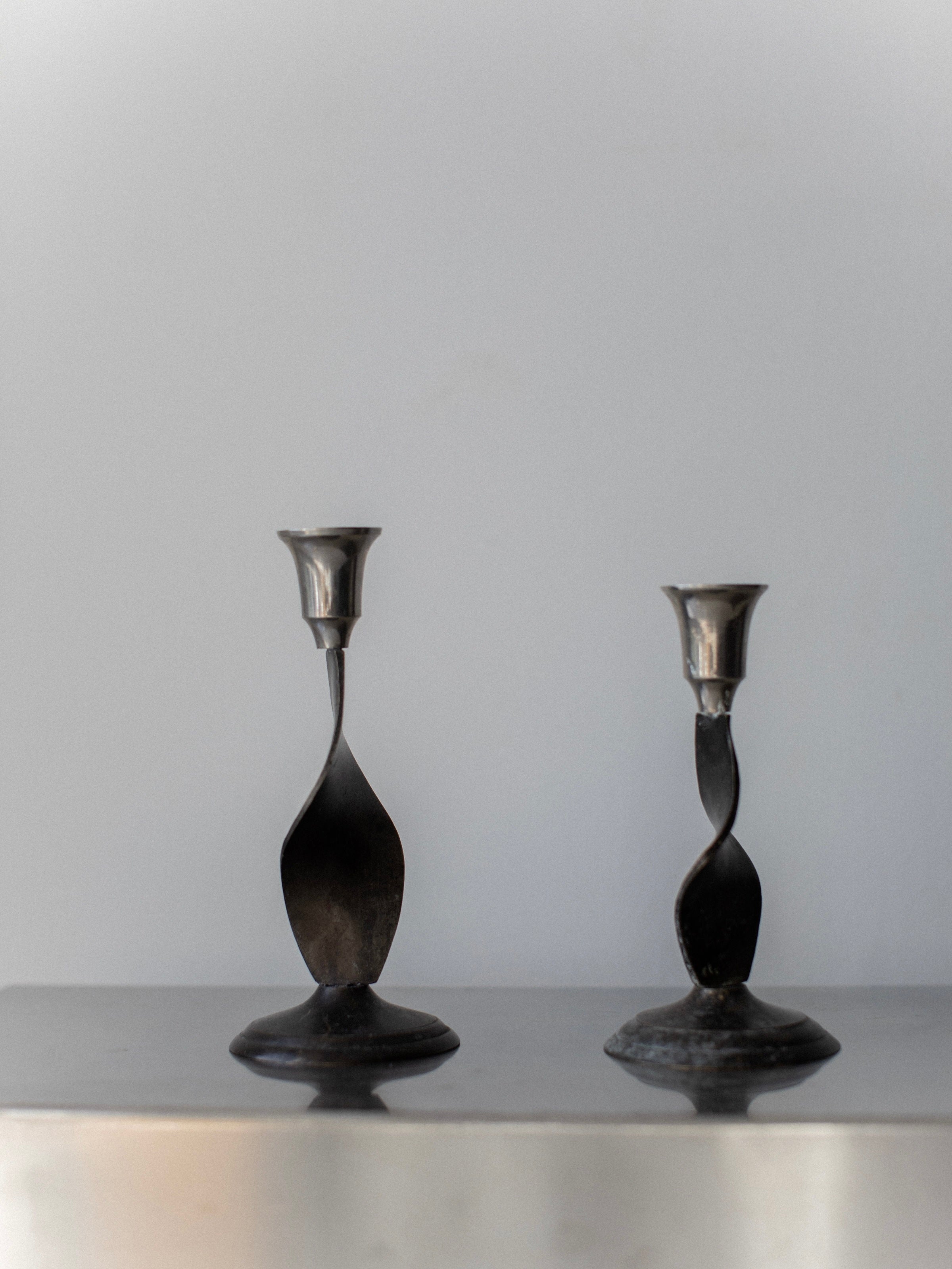 The Brutalist Candleholders by Out For Lunch, featuring an antique finish and twisted metal design, rest on a smooth, reflective surface, capturing the essence of 70's brutalist home decor against a light gray background.